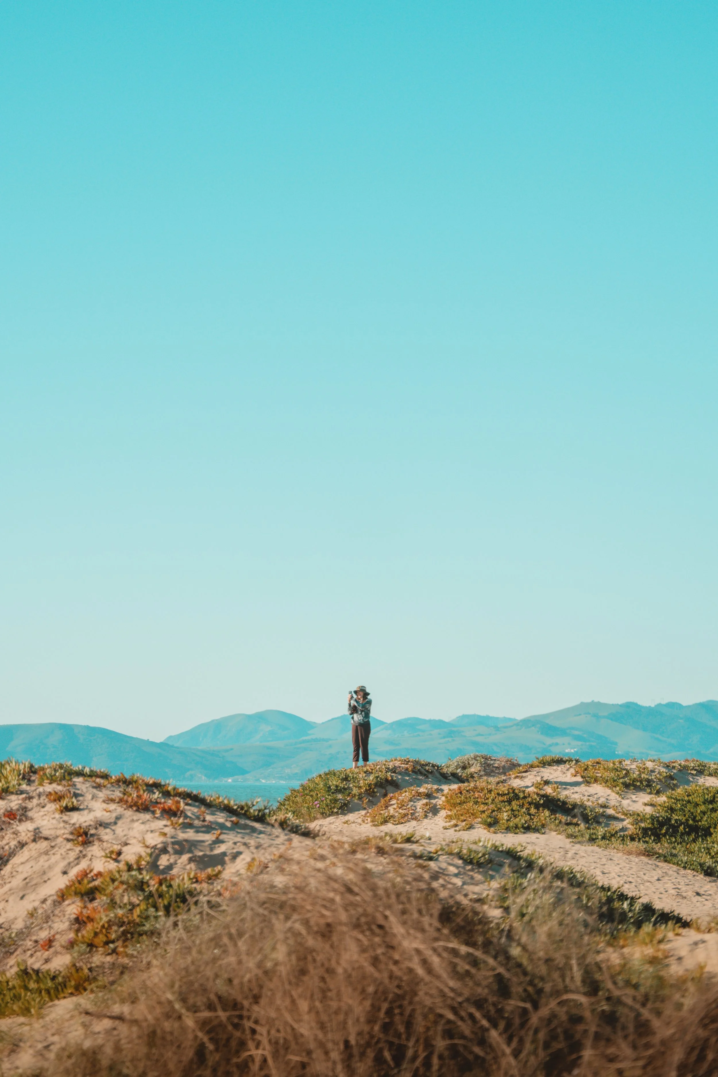 A person standing on a hilltop, taking a photo of the landscape with mountains in the background and a clear blue sky.