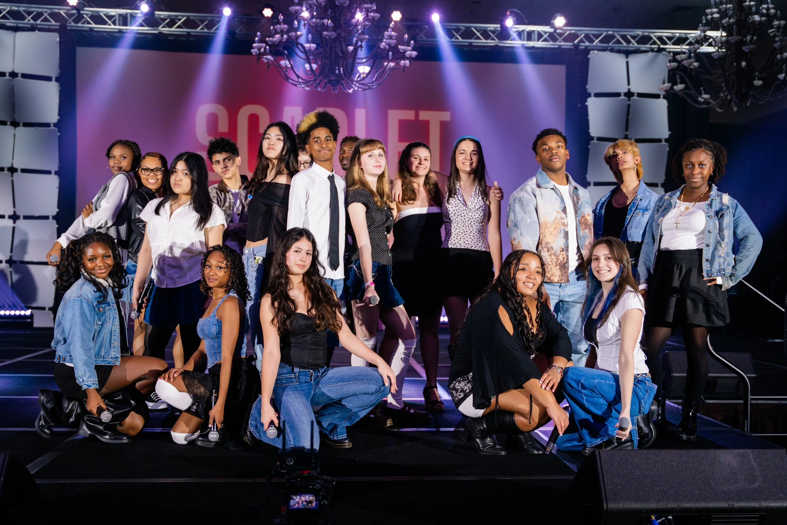 Group of diverse young people standing and kneeling on stage during an event with a large screen displaying the word 'SOLIDARITY' in the background and stage lights overhead.
