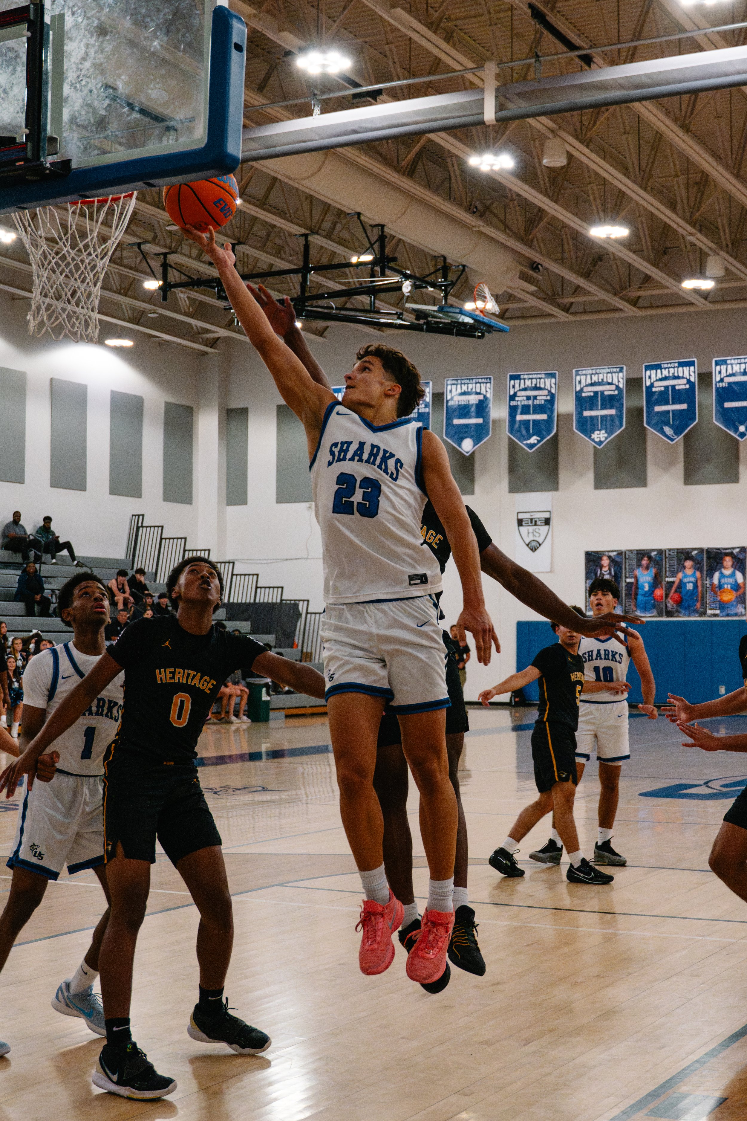 A basketball player wearing a white jersey with the team name 'SHARKS' and the number 23 is jumping to make a shot near the basket, with other players watching and reaching out around him in a gymnasium.