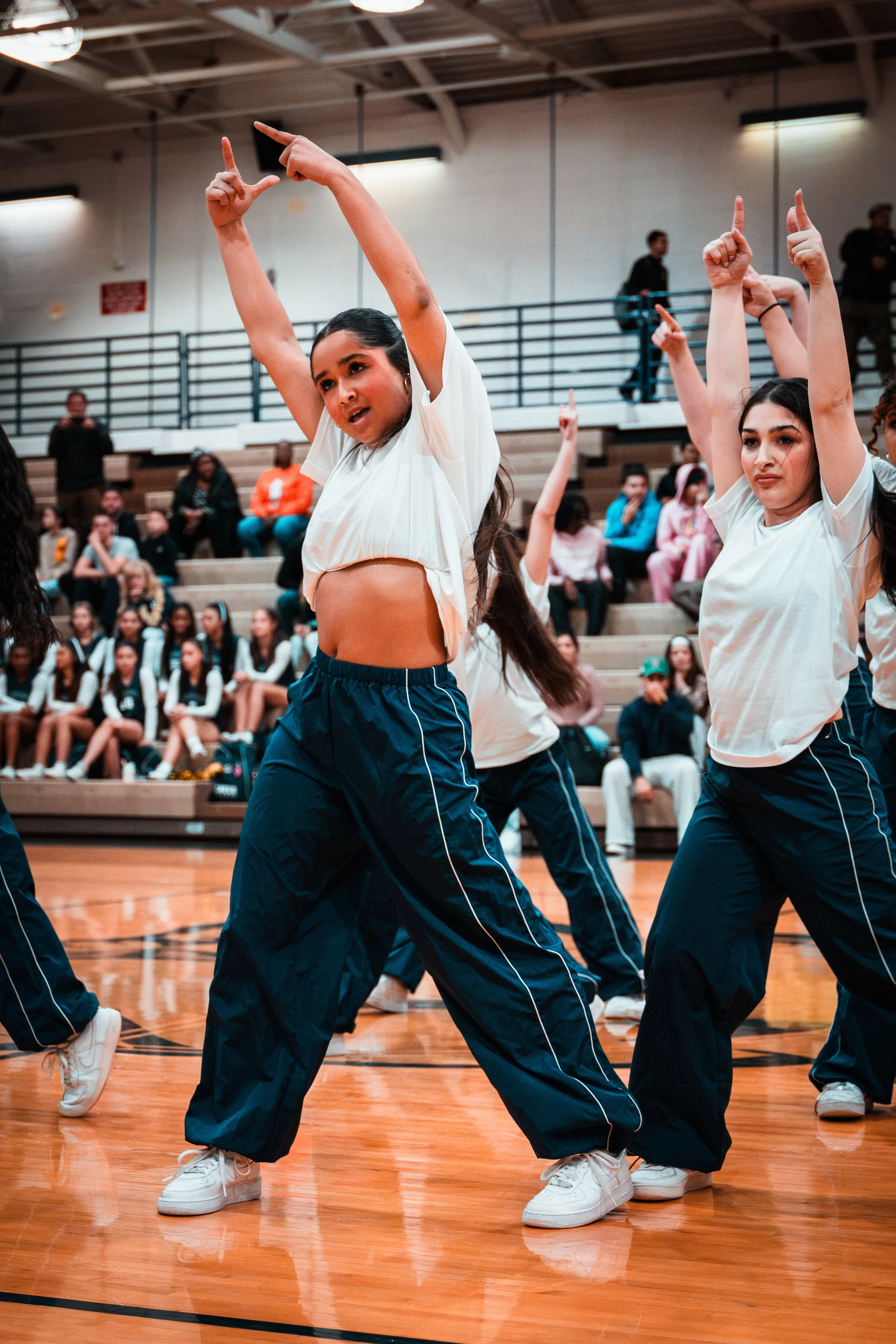 Group of young women dancing in a gymnasium, with audience seated in the background.