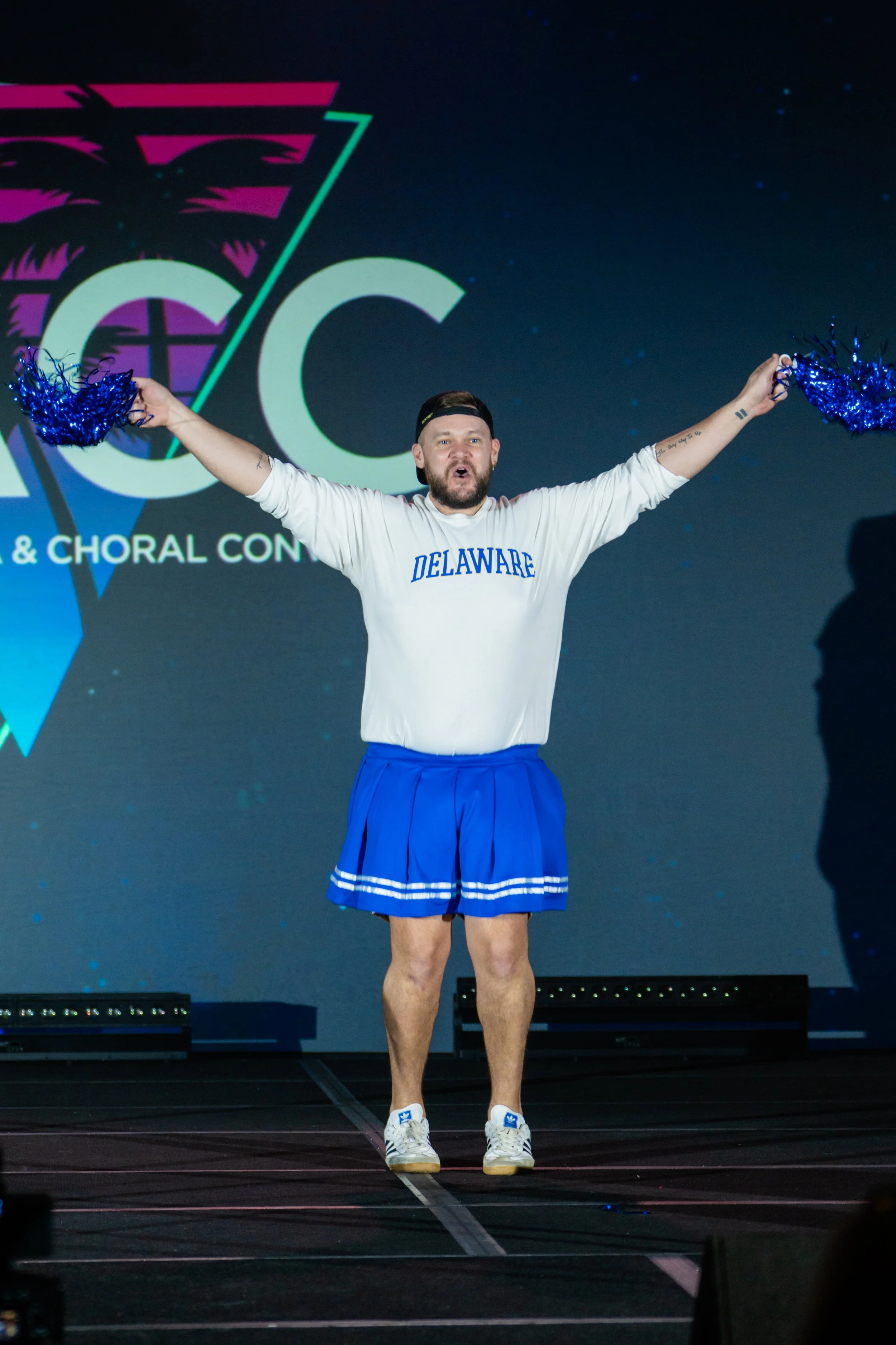 A man standing on stage with his arms raised, holding blue pom-poms in each hand, wearing a white long-sleeve shirt with 'DELAWARE' written on it, a black cap, blue cheerleading skirt, and sneakers. Behind him is a large screen with a logo and the wo
