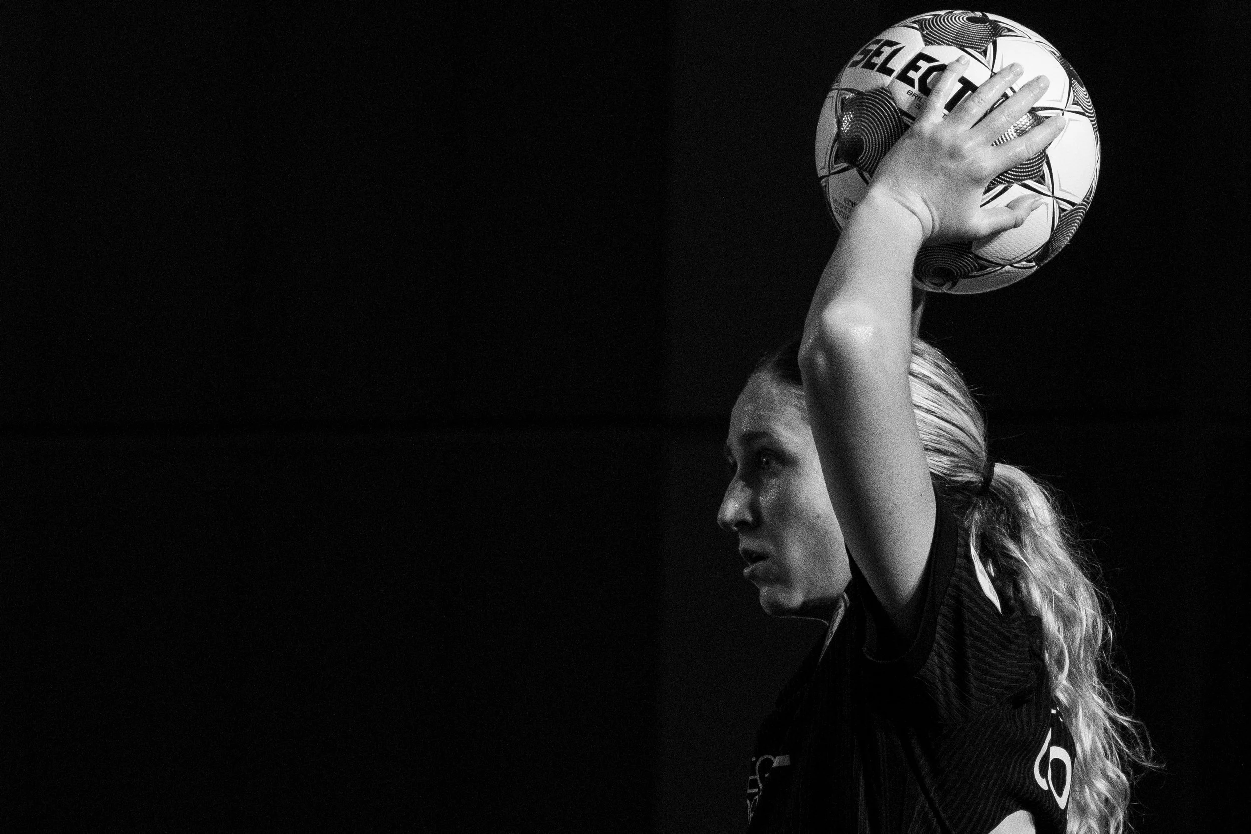 A black and white photo of a female soccer player holding a ball above her head, her face in profile, with long, curly hair, wearing a sports jersey.