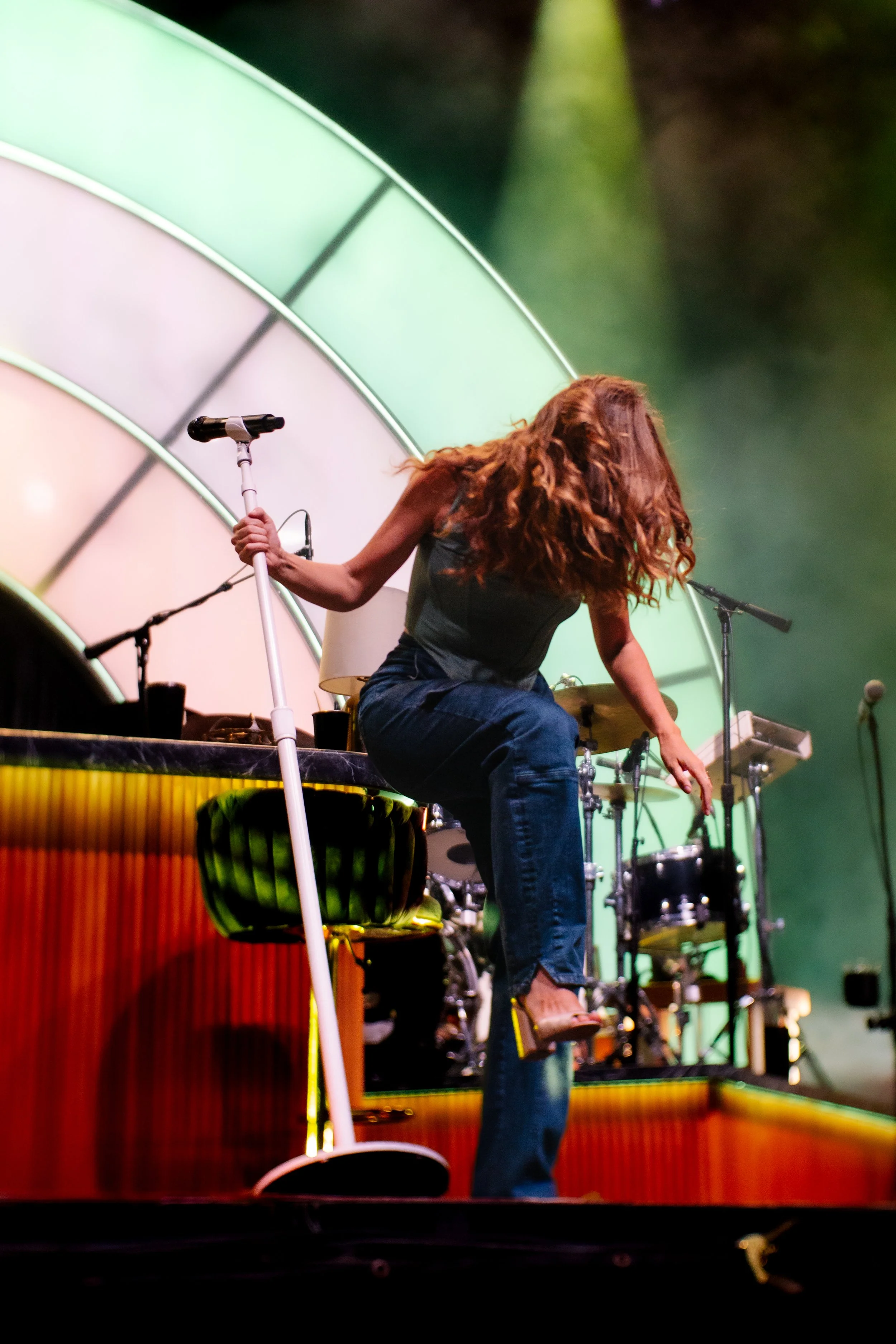 A woman with long curly hair on stage, adjusting a drum set while holding a microphone stand, with colorful stage lighting and a percussion instrument nearby.