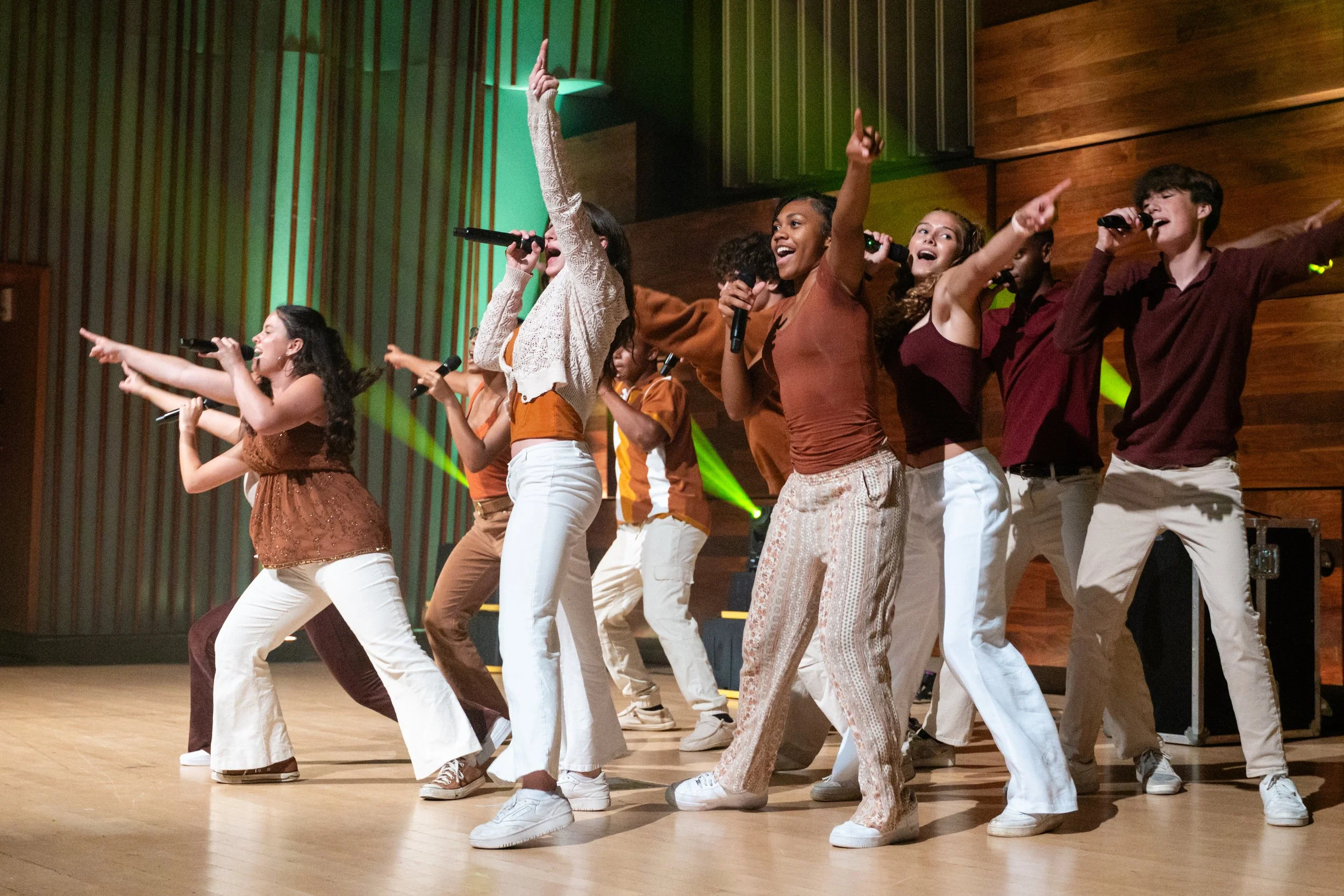 Group of young people performing on stage with microphones, singing and dancing enthusiastically, decorated with wood paneling and green stage lighting.