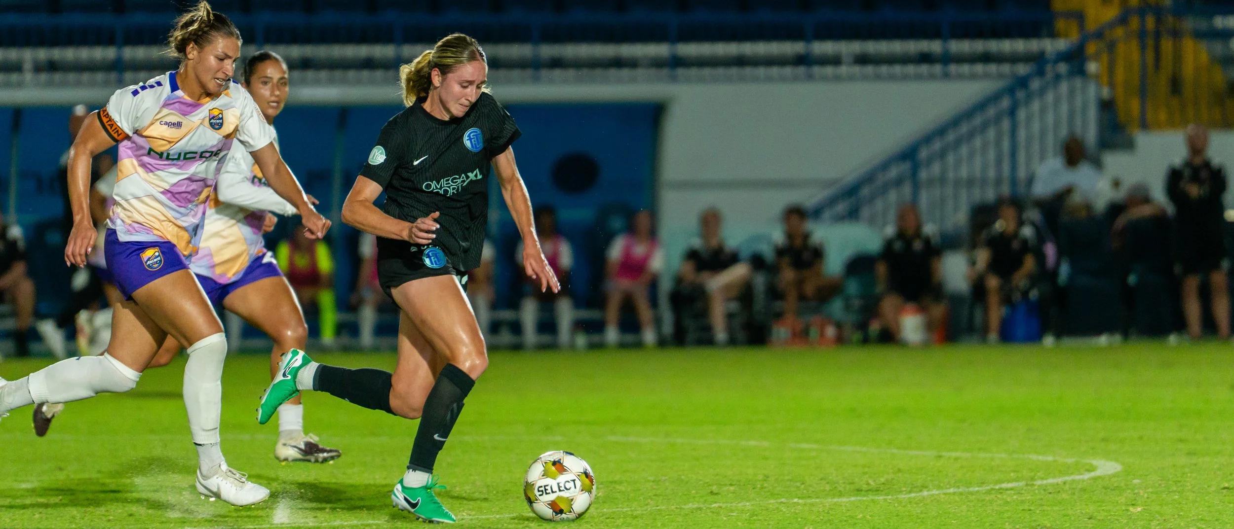 Women playing soccer on a green field under stadium lights, with spectators in the background.