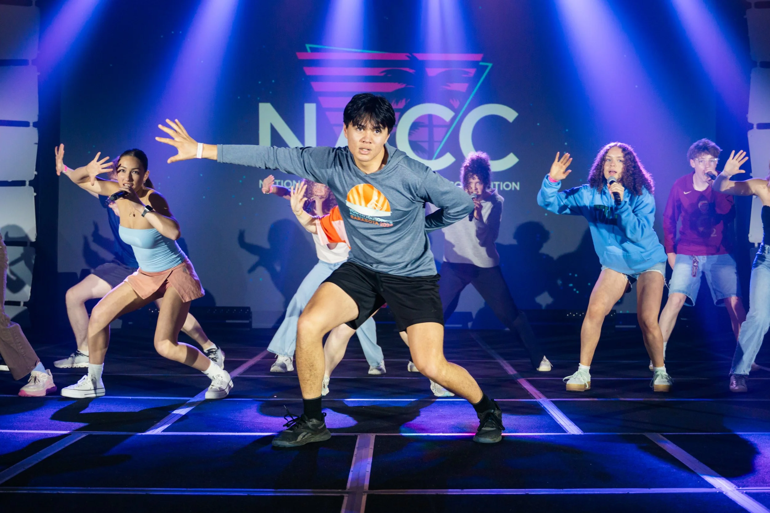 Young man dancing on stage during a performance, surrounded by backup dancers, with a neon-lit "NEC" logo in the background.