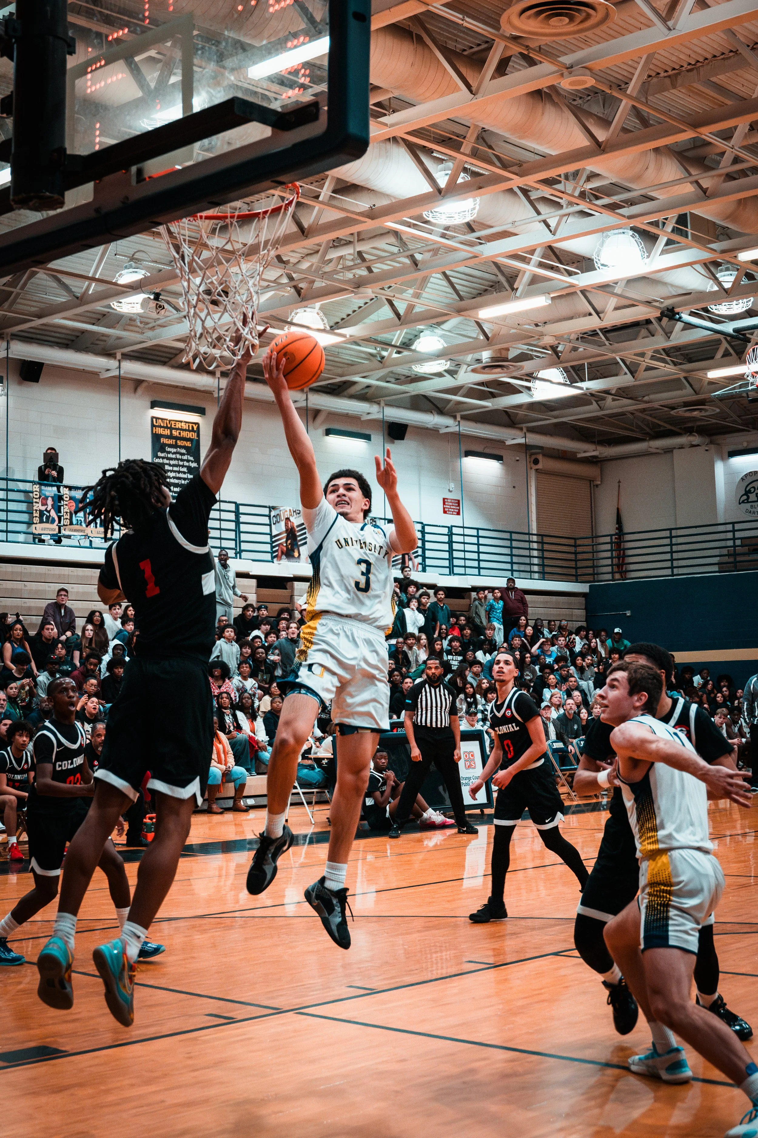 A basketball game in a gymnasium with players competing near the hoop. A player in a dark uniform is attempting to block a shot by a player in a white uniform with number 3, who is jumping toward the basket. The gym is filled with spectators watching