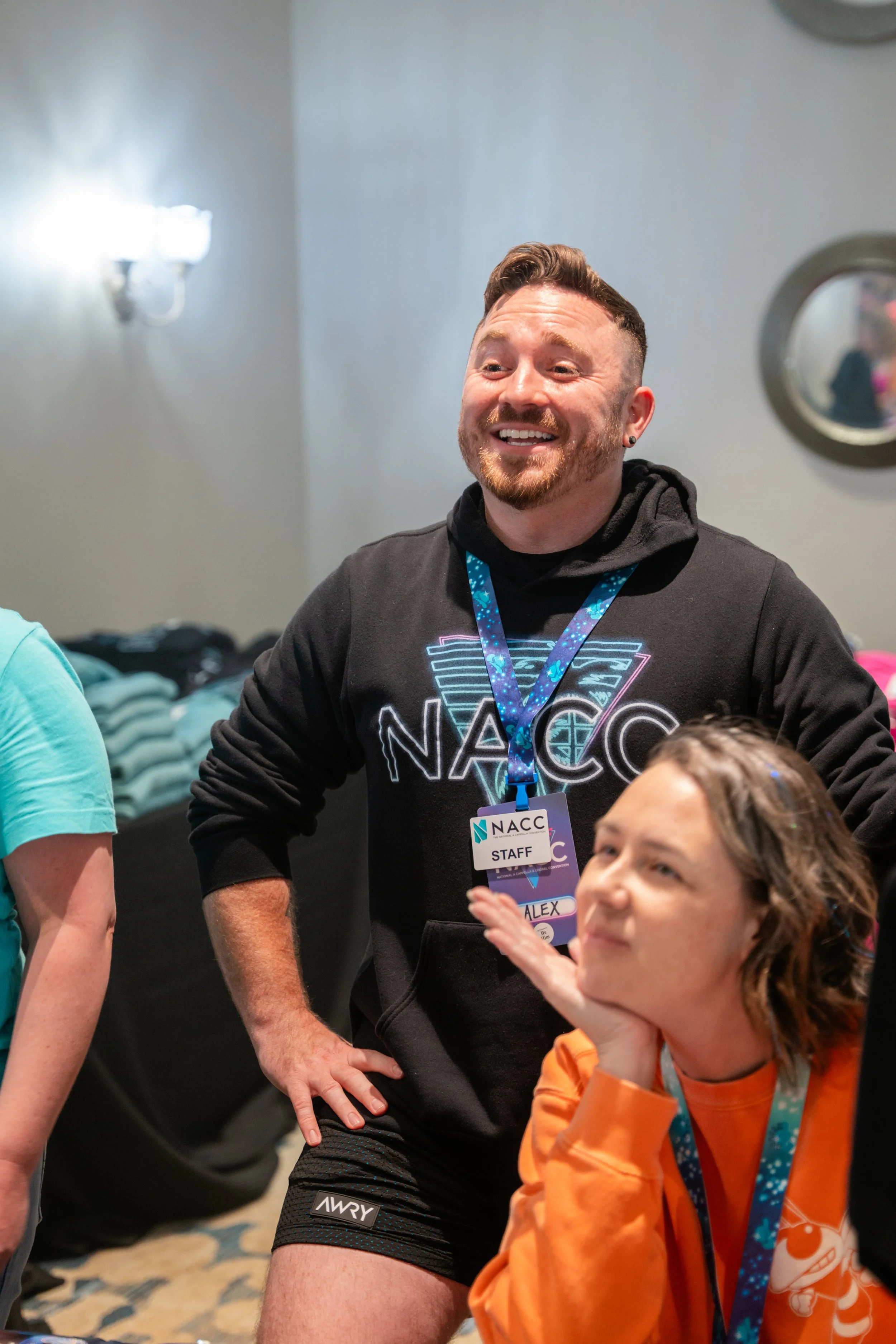 A man with short brown hair, a beard, and earrings wearing a black NASA hoodie and a staff badge, smiling and talking to children at an indoor event.