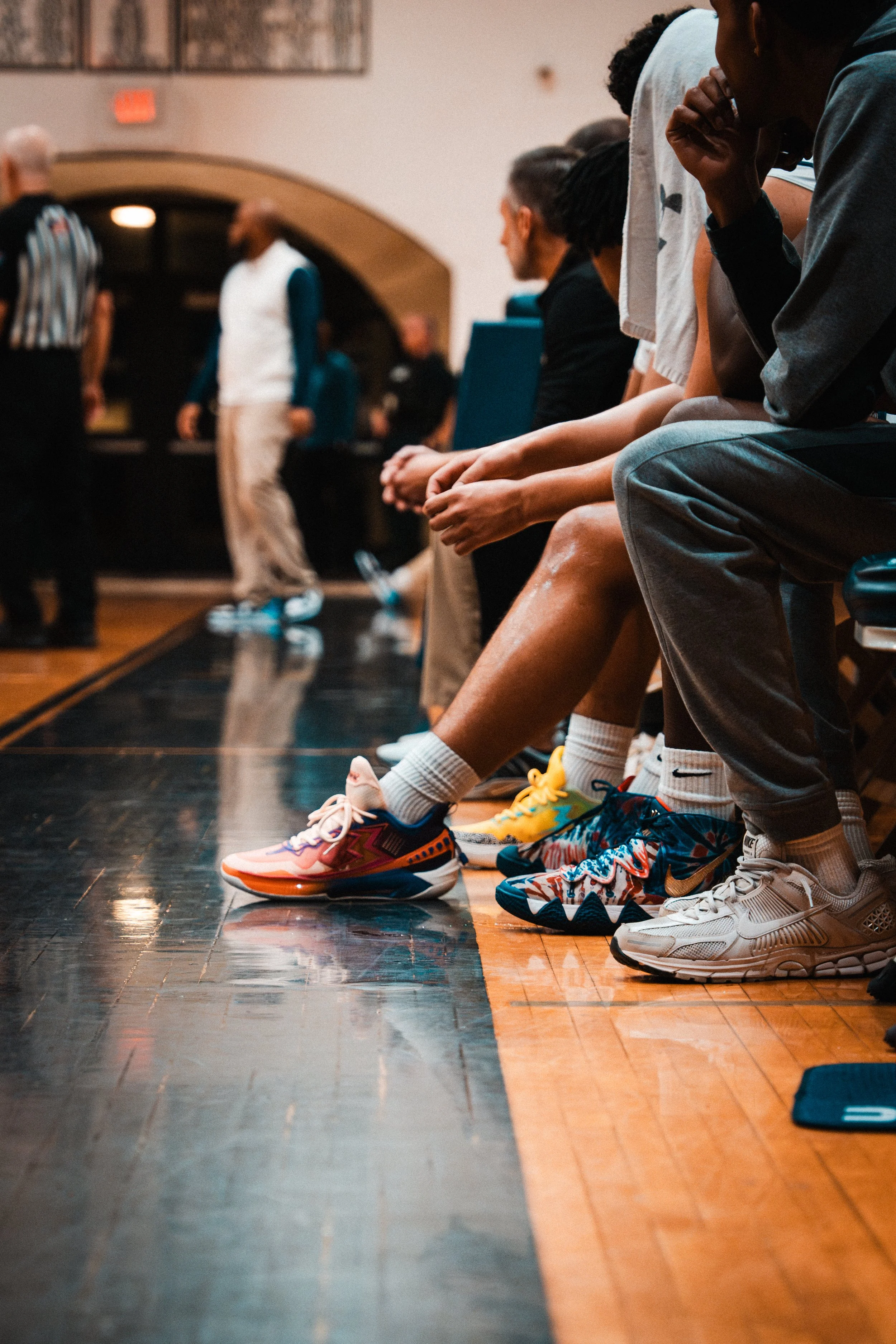 Close-up of a row of basketball players sitting on a bench during a game, with their legs and colorful athletic shoes prominently visible.