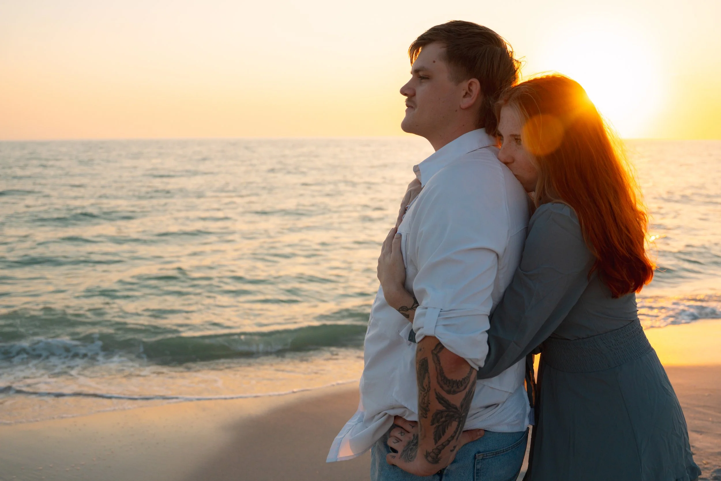 A couple embraces on the beach at sunset, with the woman hugging the man from behind and the ocean in the background.