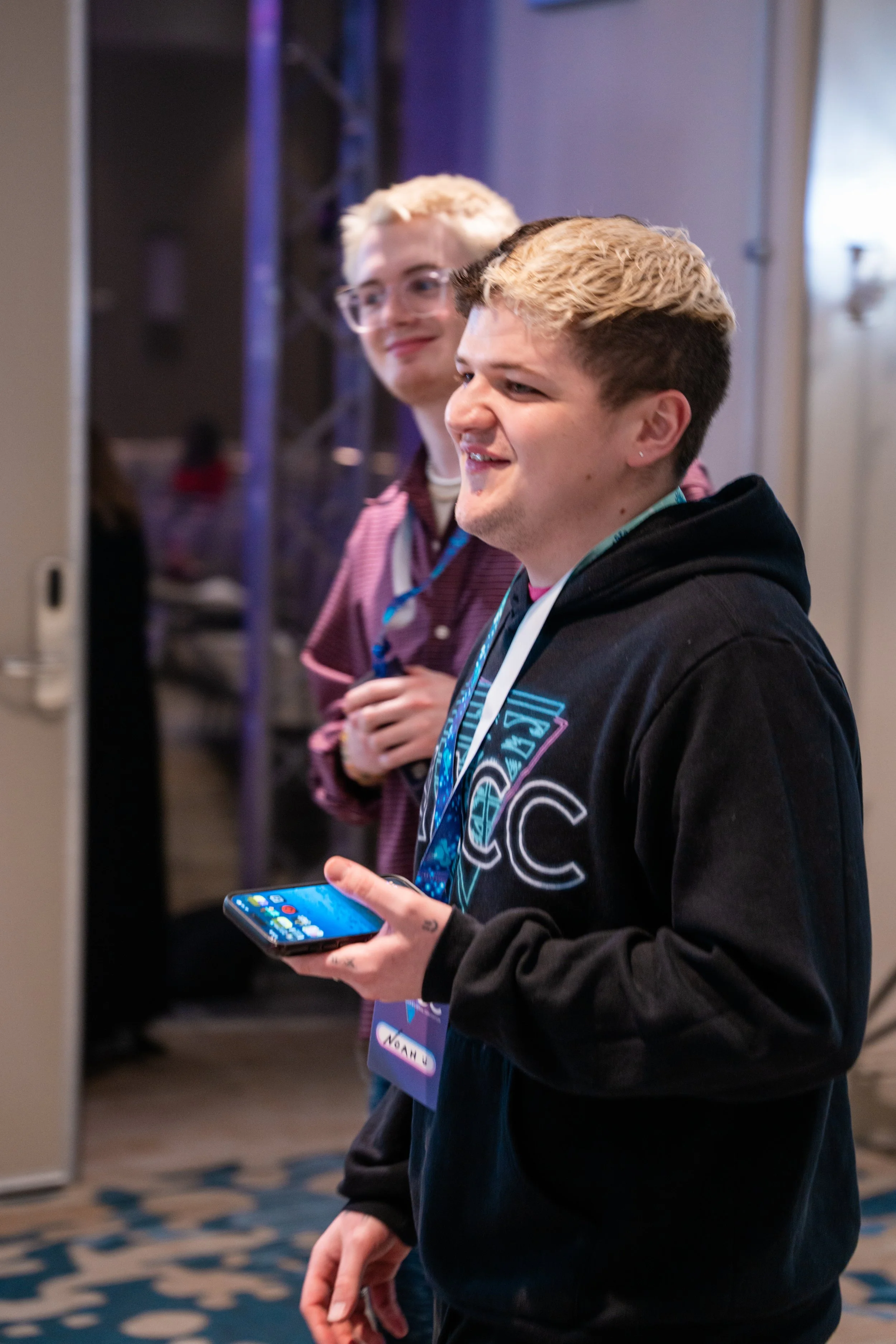Two young men standing at an indoor event, one holding a smartphone, both wearing lanyards and smiling.