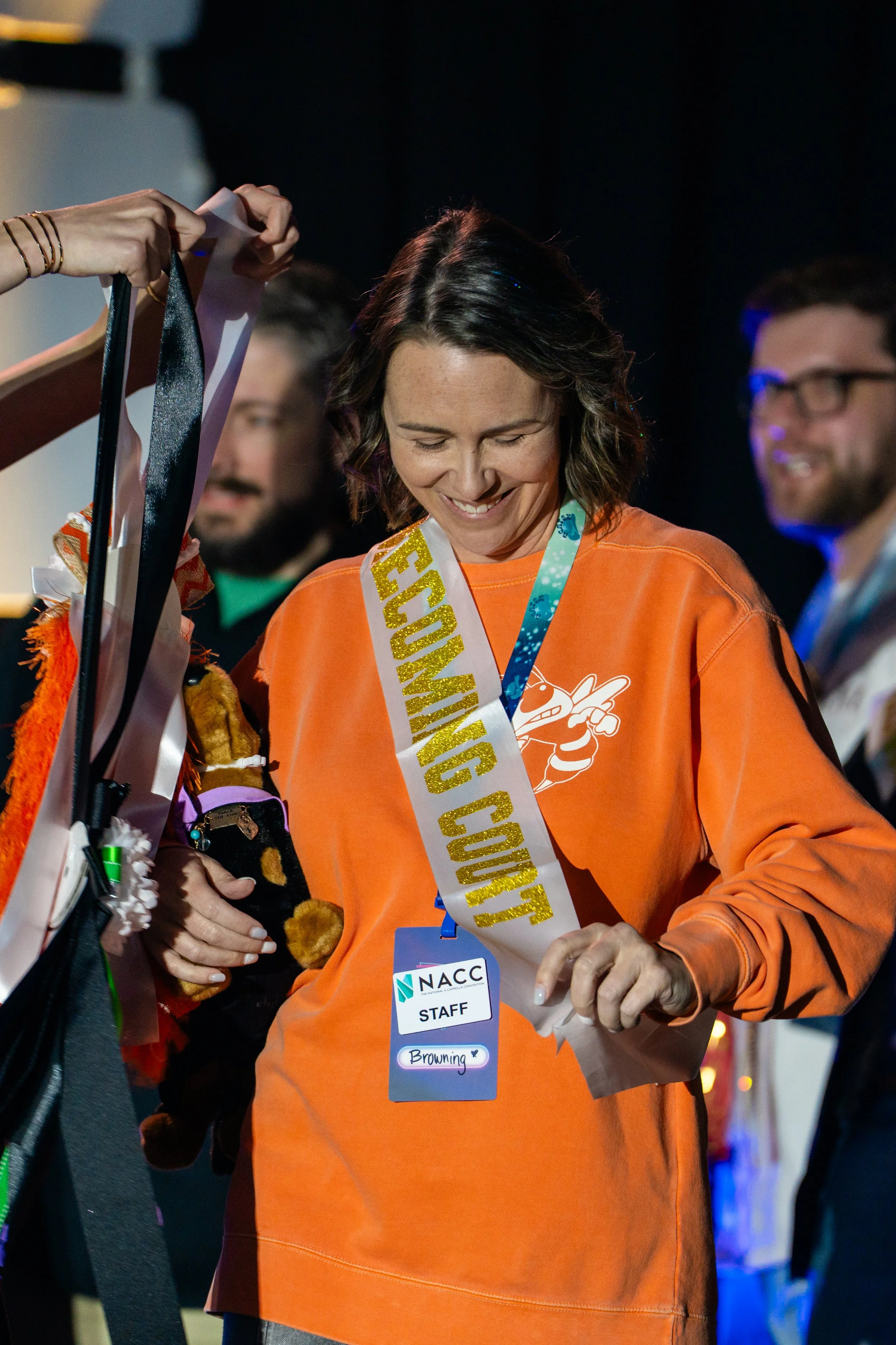 A woman wearing orange sweatshirt and a sash that says 'ECONOMIC CURE' is smiling while standing with others. She has a staff badge with the name 'Browning' and a blue lanyard with the 'NACC' logo. She appears to be at an event.