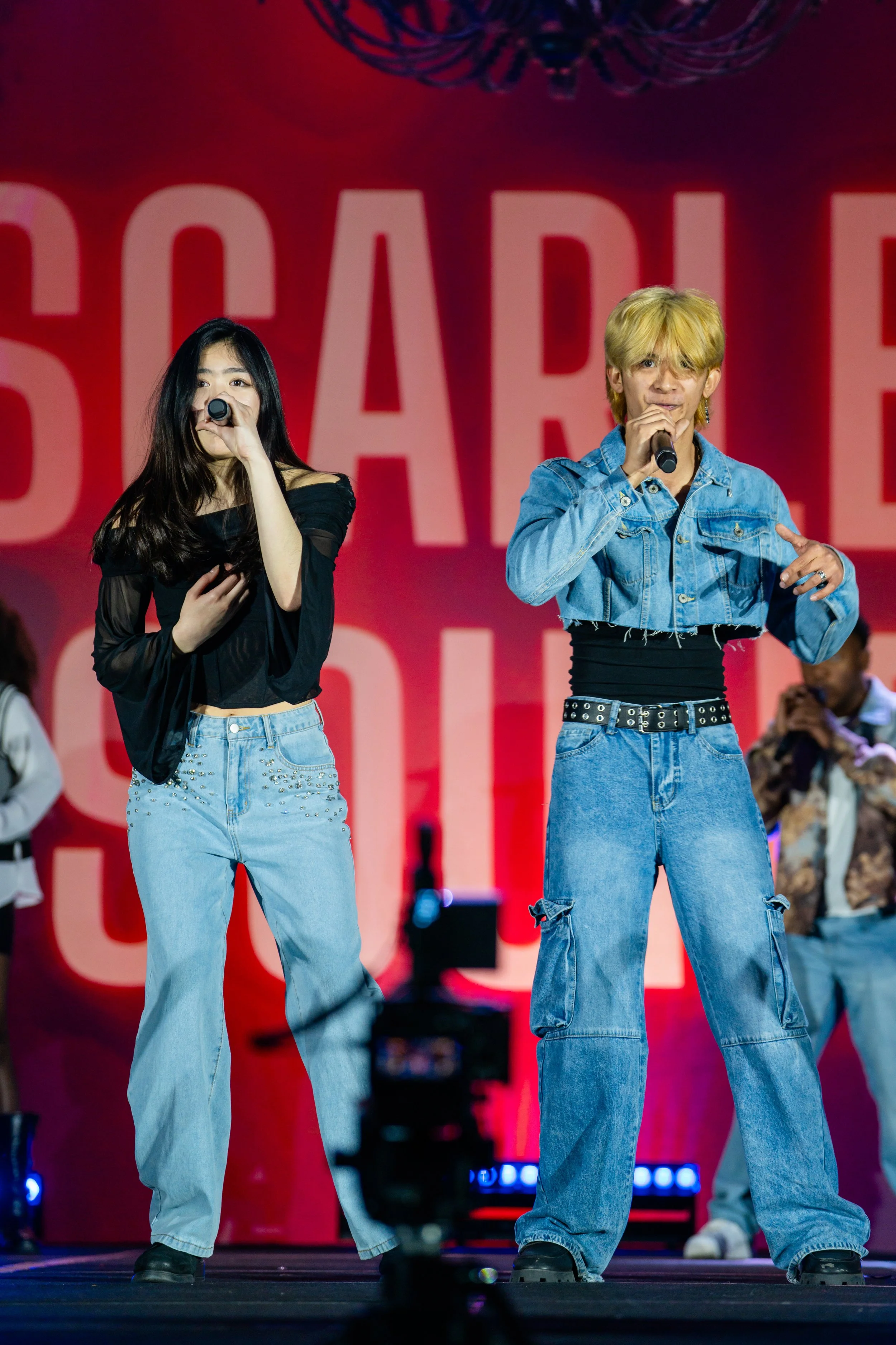 Two young performers on stage, one woman with long dark hair wearing a black off-the-shoulder top and light blue jeans with embellishments, and one man with blond hair in a denim jacket and baggy jeans, both holding microphones and singing. The backg