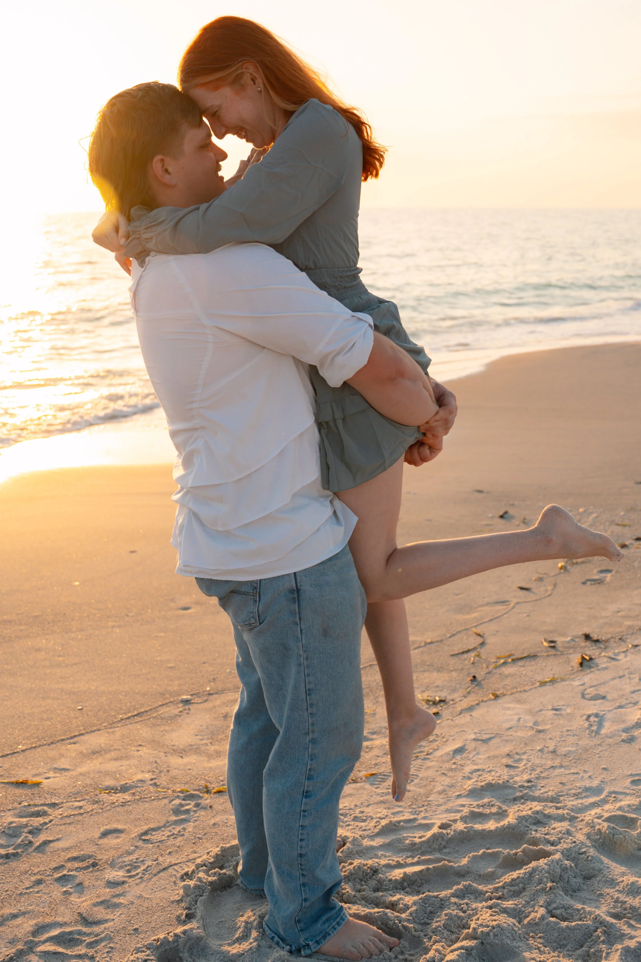 A man lifting a woman, both smiling and touching foreheads, on a beach during sunset.