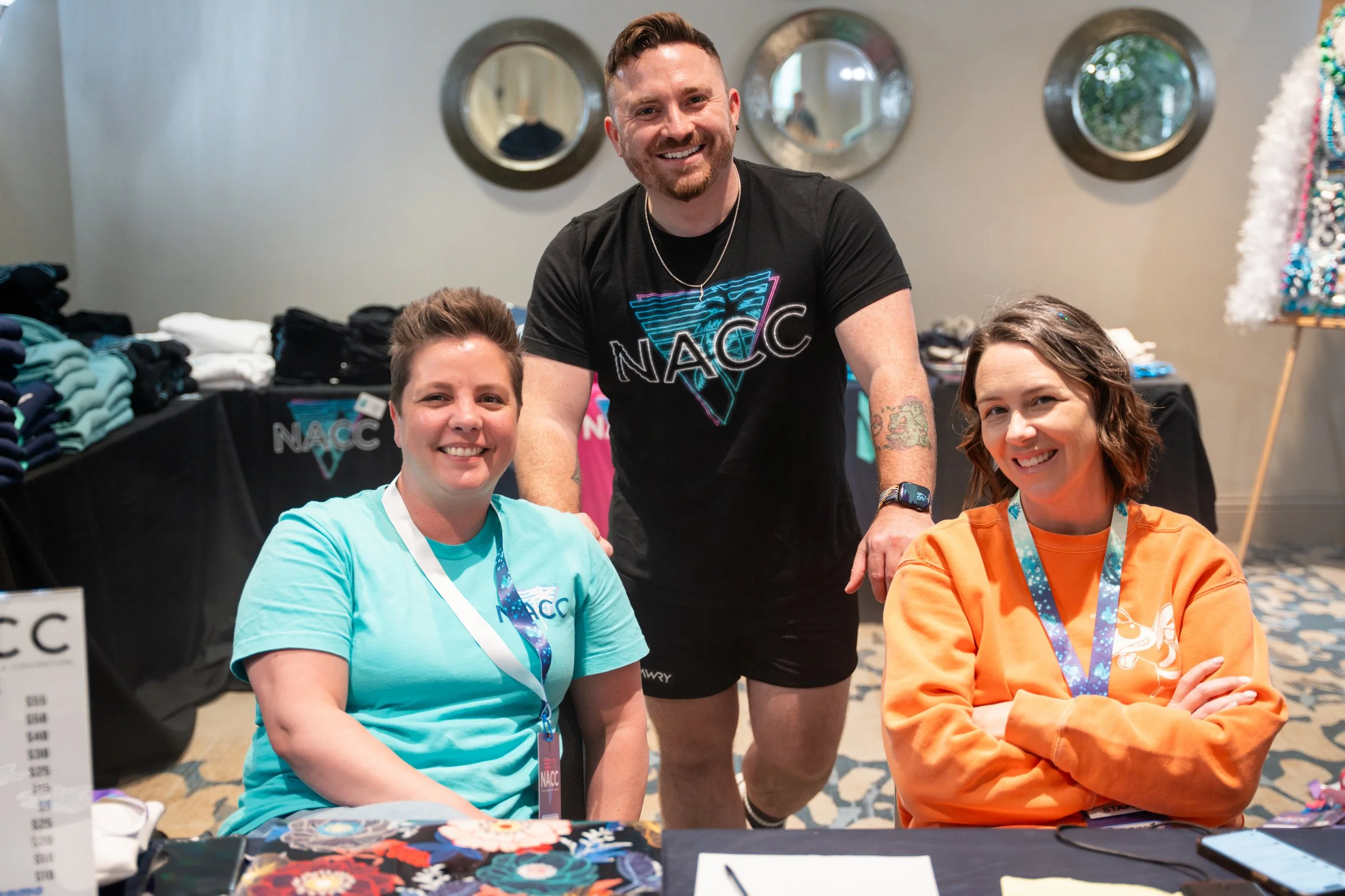 Three people at a booth with cloth and merchandise. The woman on the left wears a light blue shirt and white lanyard, the woman on the right wears an orange sweatshirt, and the man in the center stands behind them with a black T-shirt, smiling.