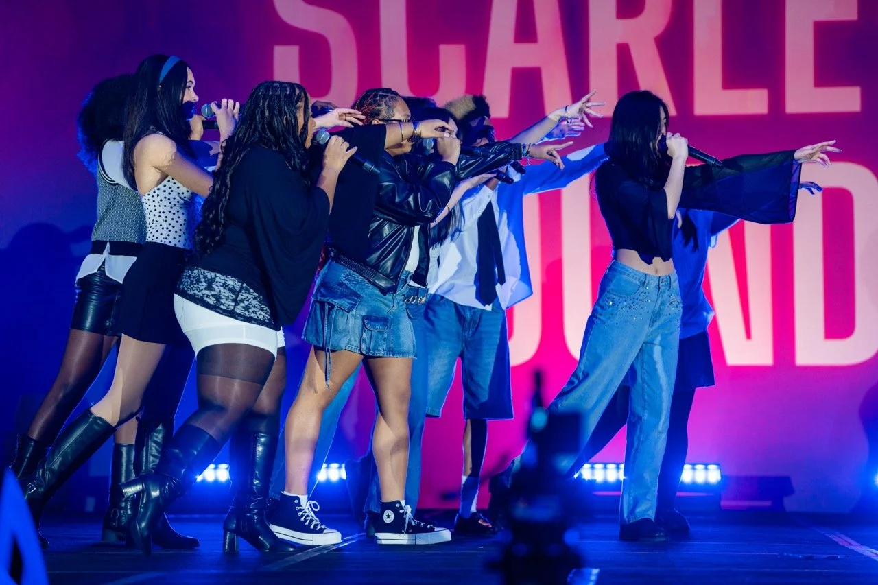 A group of young women performing on stage, some holding microphones and reaching out, with a pink and purple background that has the word 'SHAREL' partially visible.