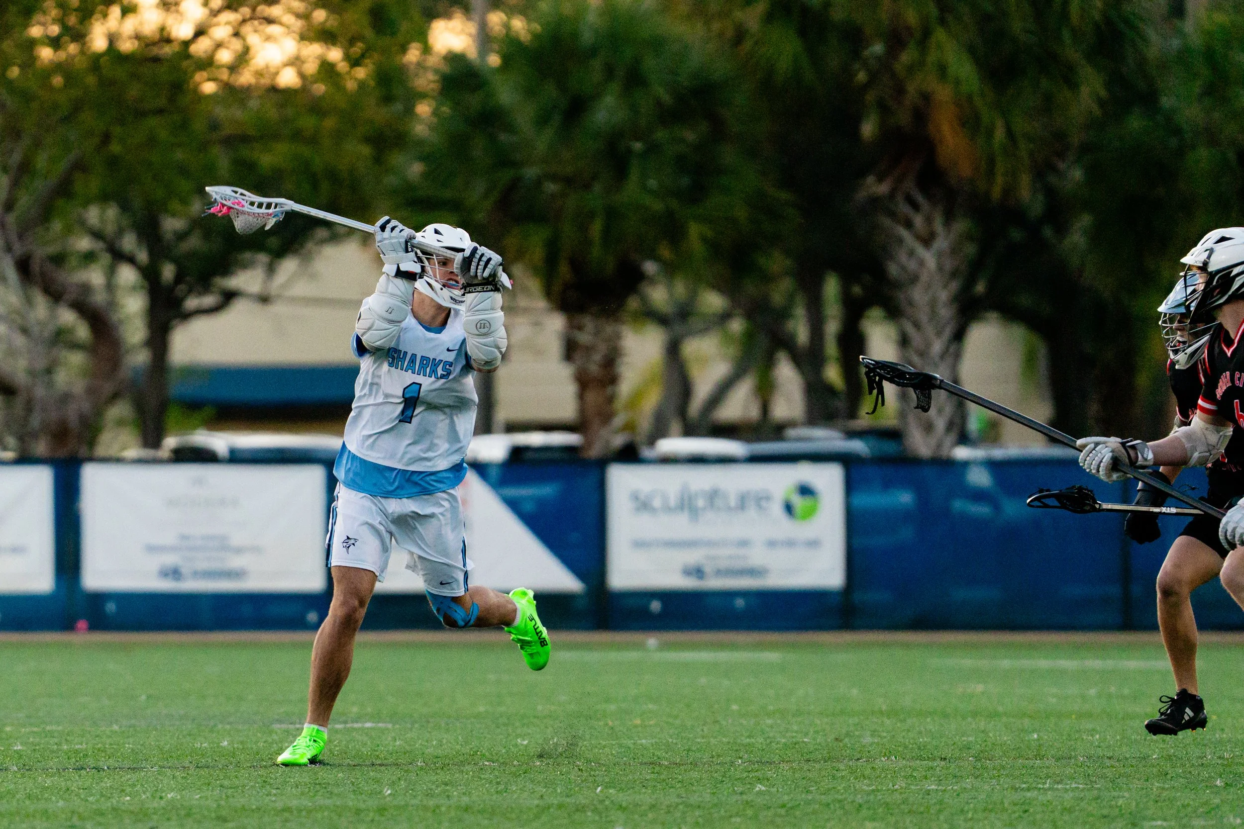 A lacrosse player in a white jersey with blue accents and the word 'SHARKS' on front, wearing protective gear, is catching a ball with his lacrosse stick during a game on a grassy field. Another player in black jersey with red accents is also in the 