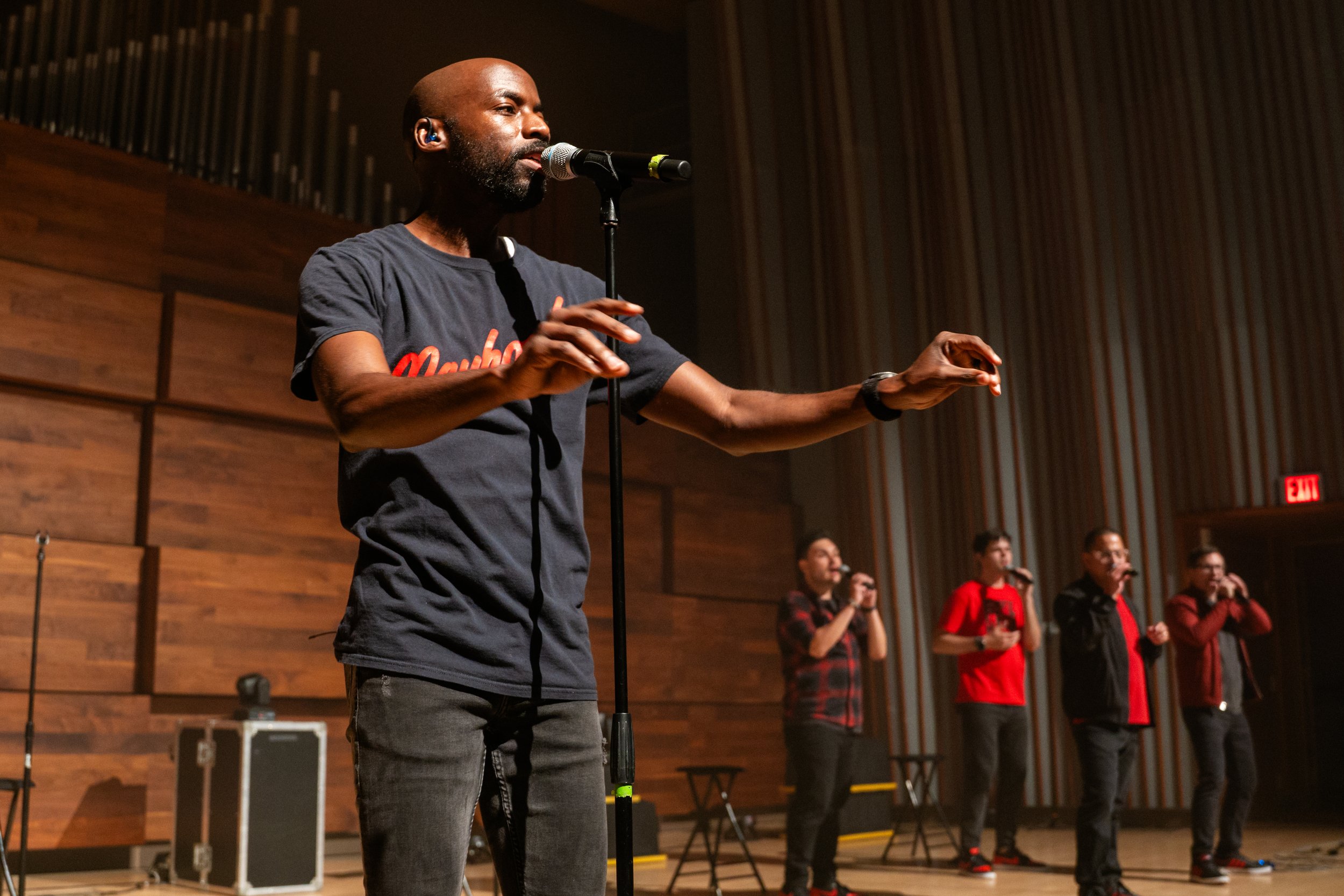 A man with a beard is singing into a microphone on a stage, with four other men in the background holding microphones. The stage has wooden walls and vertical striped panels, with musical equipment nearby.