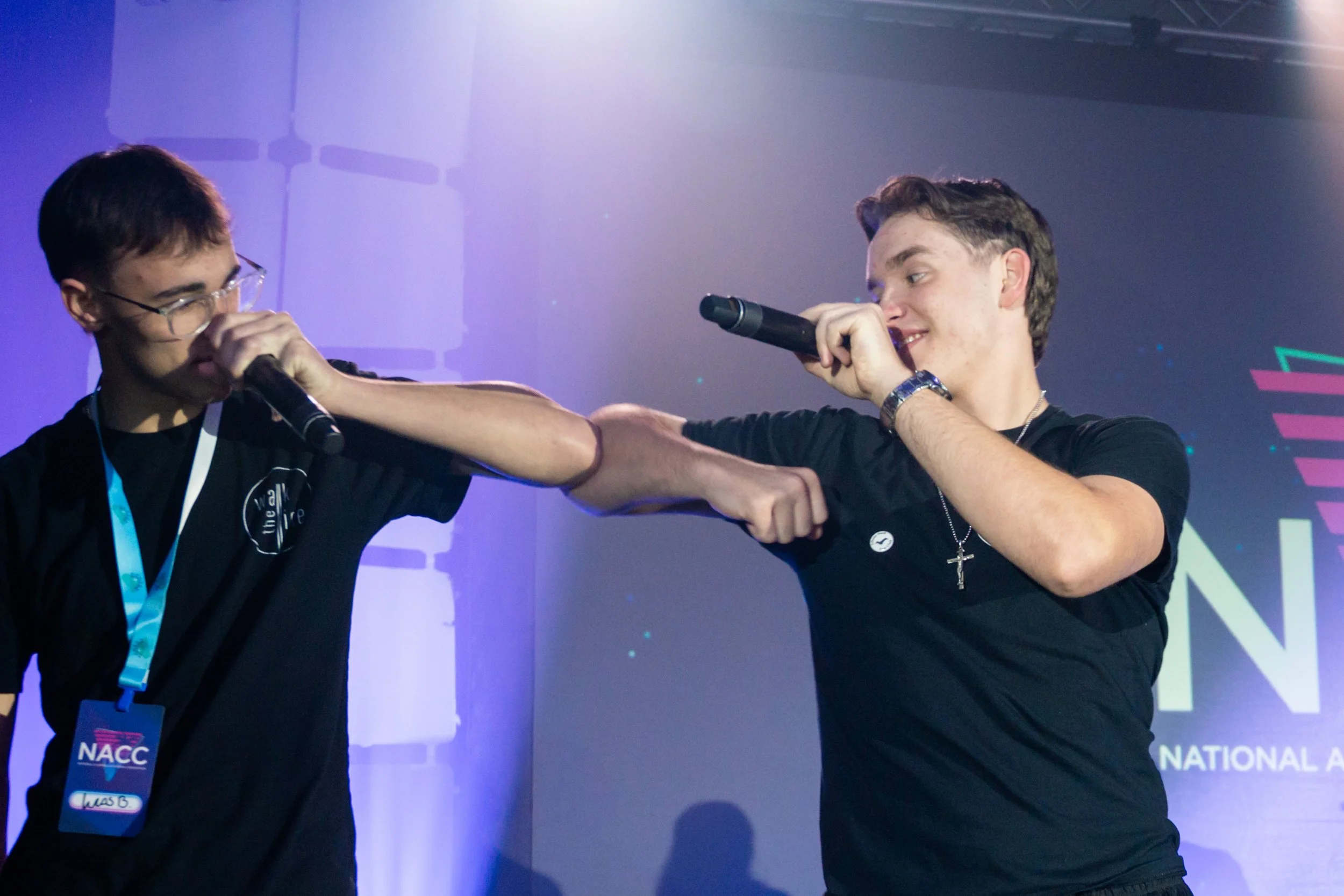 Two young men on stage engaging in a friendly fist bump, one wearing glasses and the other holding a microphone, with a colorful background and event badges.