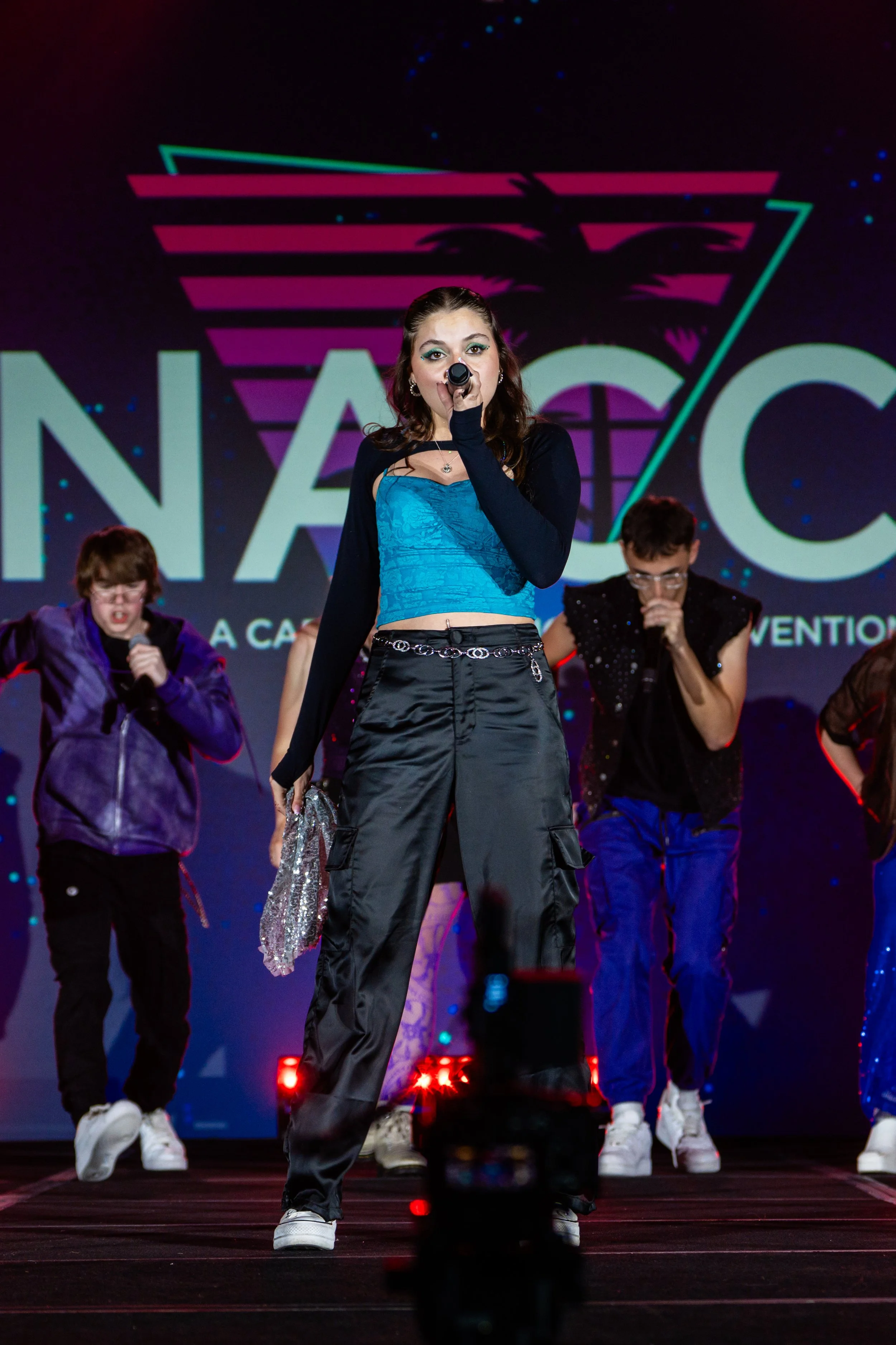 A young woman in a blue crop top, black cargo pants, with chain accessories, performing on stage with backup dancers in colorful outfits. The background features a logo with a palm tree silhouette and geometric lines.