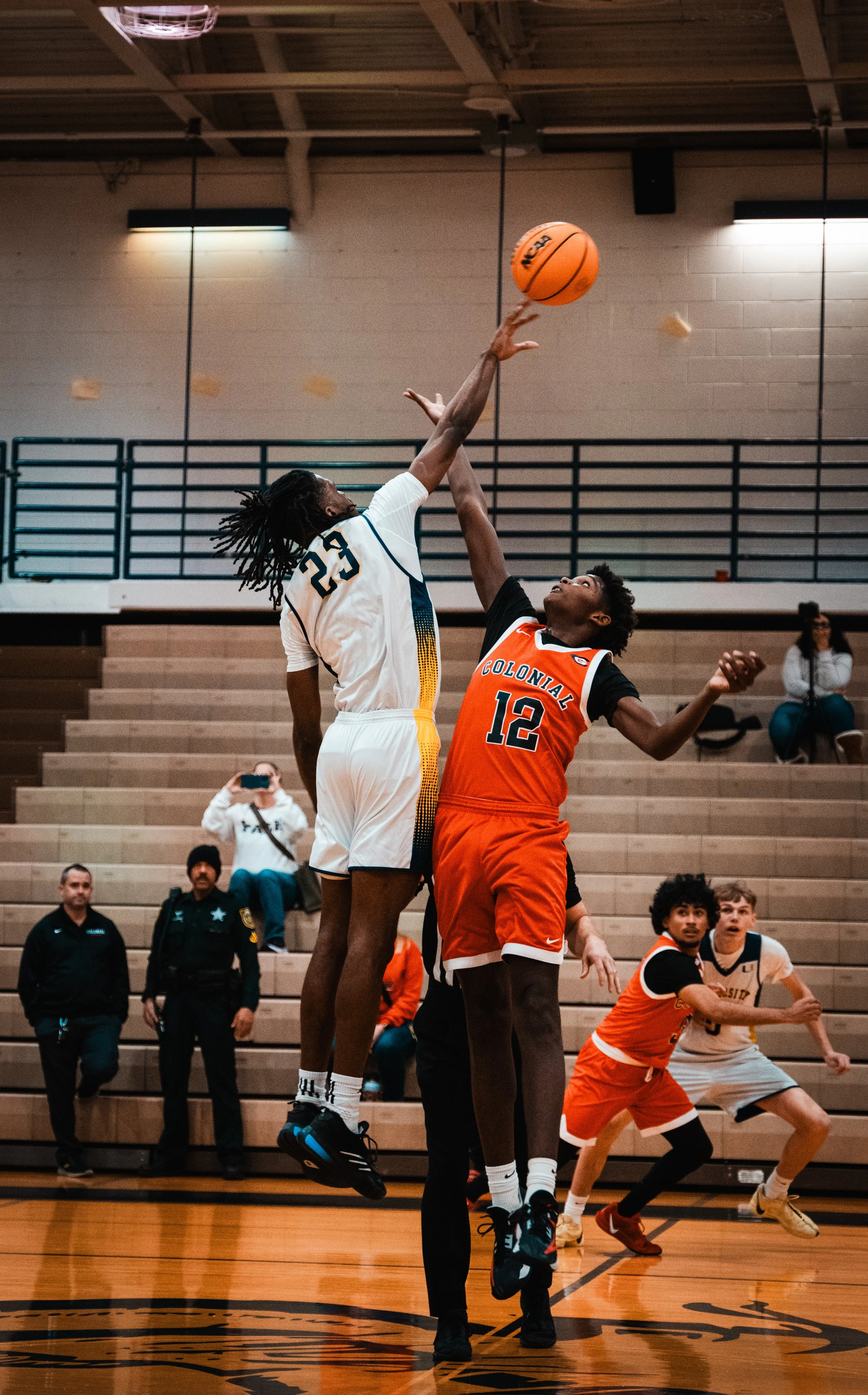 Two basketball players, one in a white jersey and one in an orange jersey, are jumping for a ball during a game, with the player in white reaching high to block the shot. There are spectators and officials on the court and in the stands.