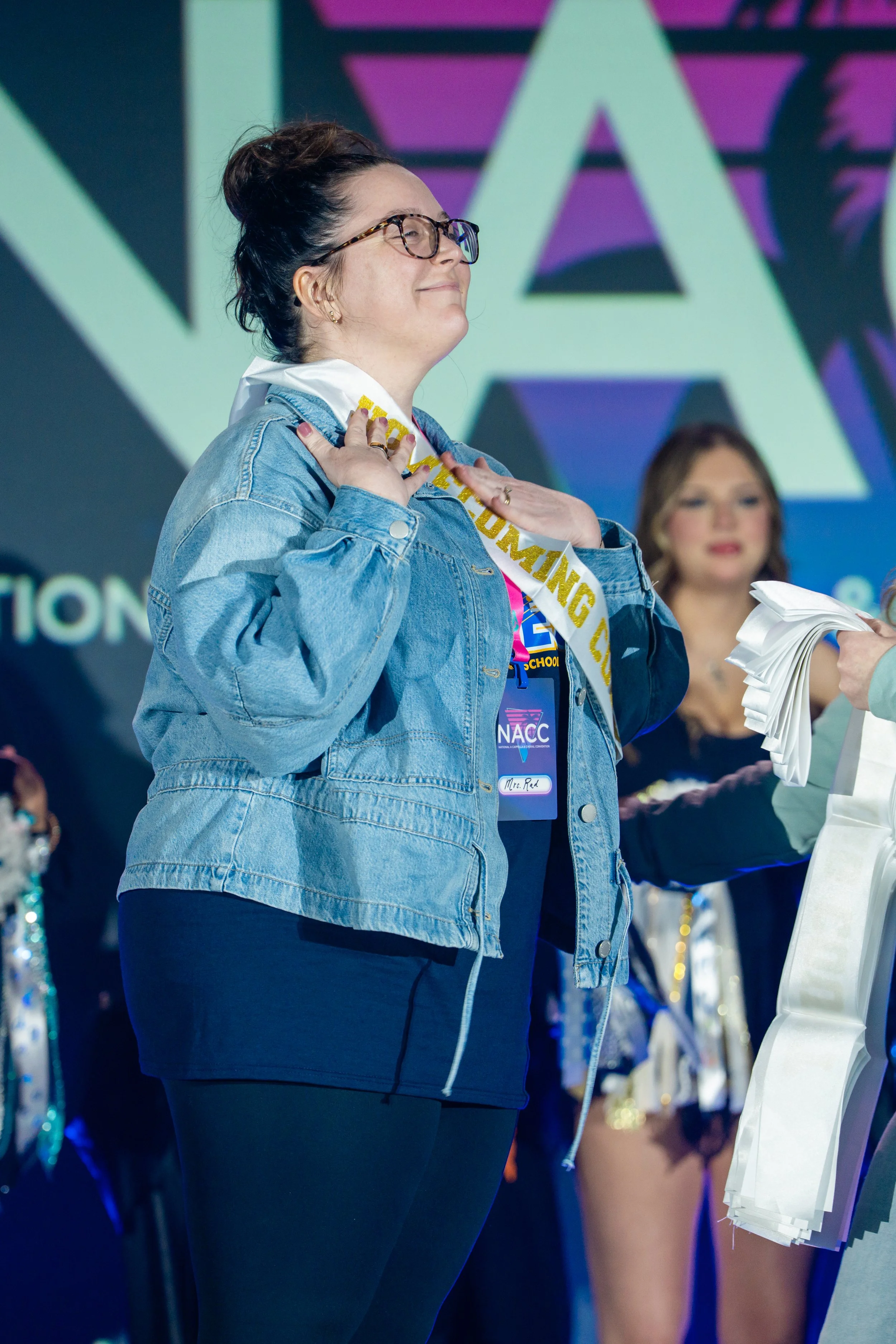 A woman wearing glasses, a denim jacket, and a sash that reads 'Graduating'. She is smiling and placing her hands on her chest, standing on stage with other women in the background at an event.
