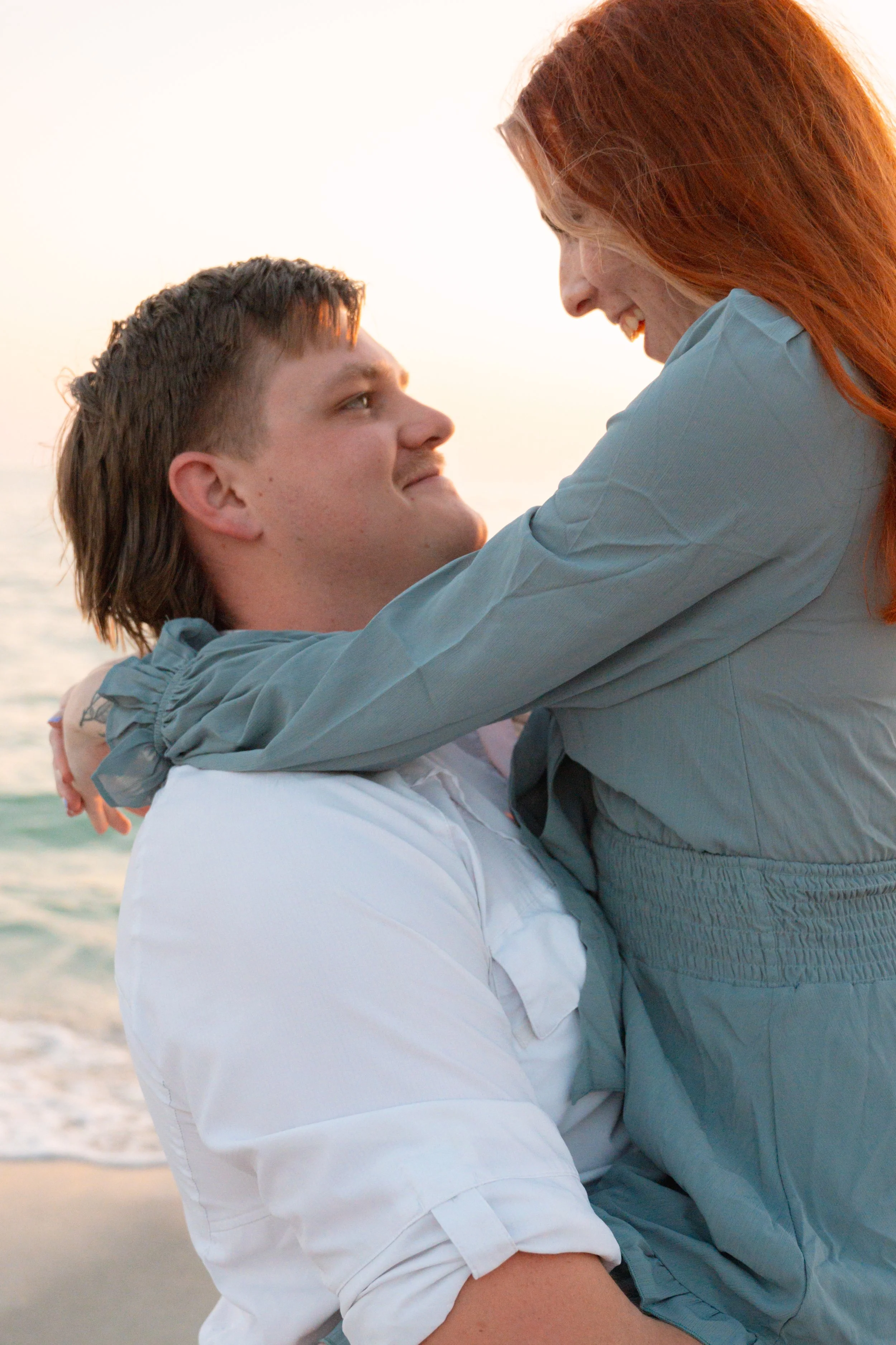 A couple at the beach, with the woman lifted in the man's arms, both smiling and enjoying each other's company during sunset.