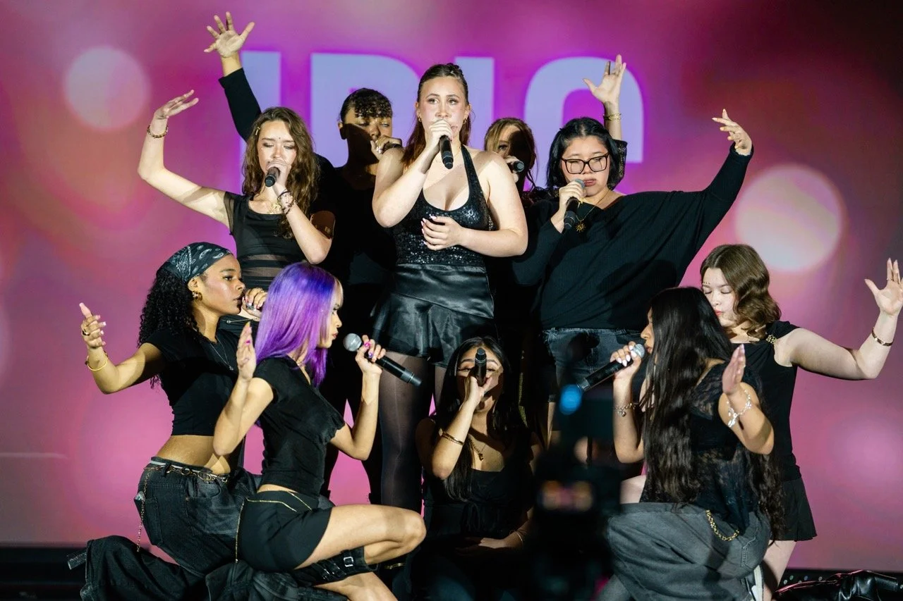 Group of young women singing and performing on stage with pink and purple lighting, some kneeling and some standing, holding microphones and raising hands, dressed in black clothing.