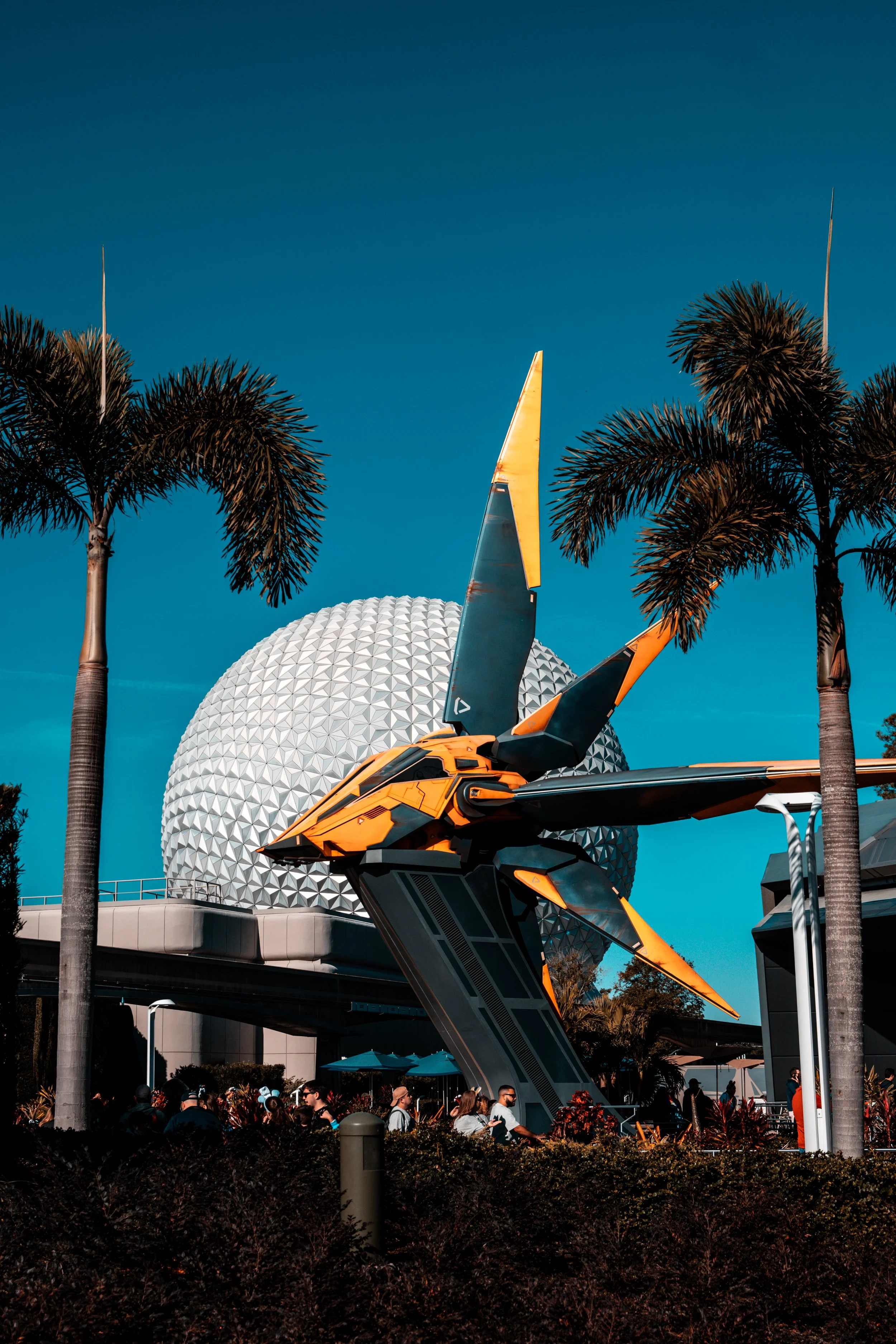 A large orange and black spaceship sculpture at Epcot theme park, with a geodesic sphere in the background and palm trees framing the scene.