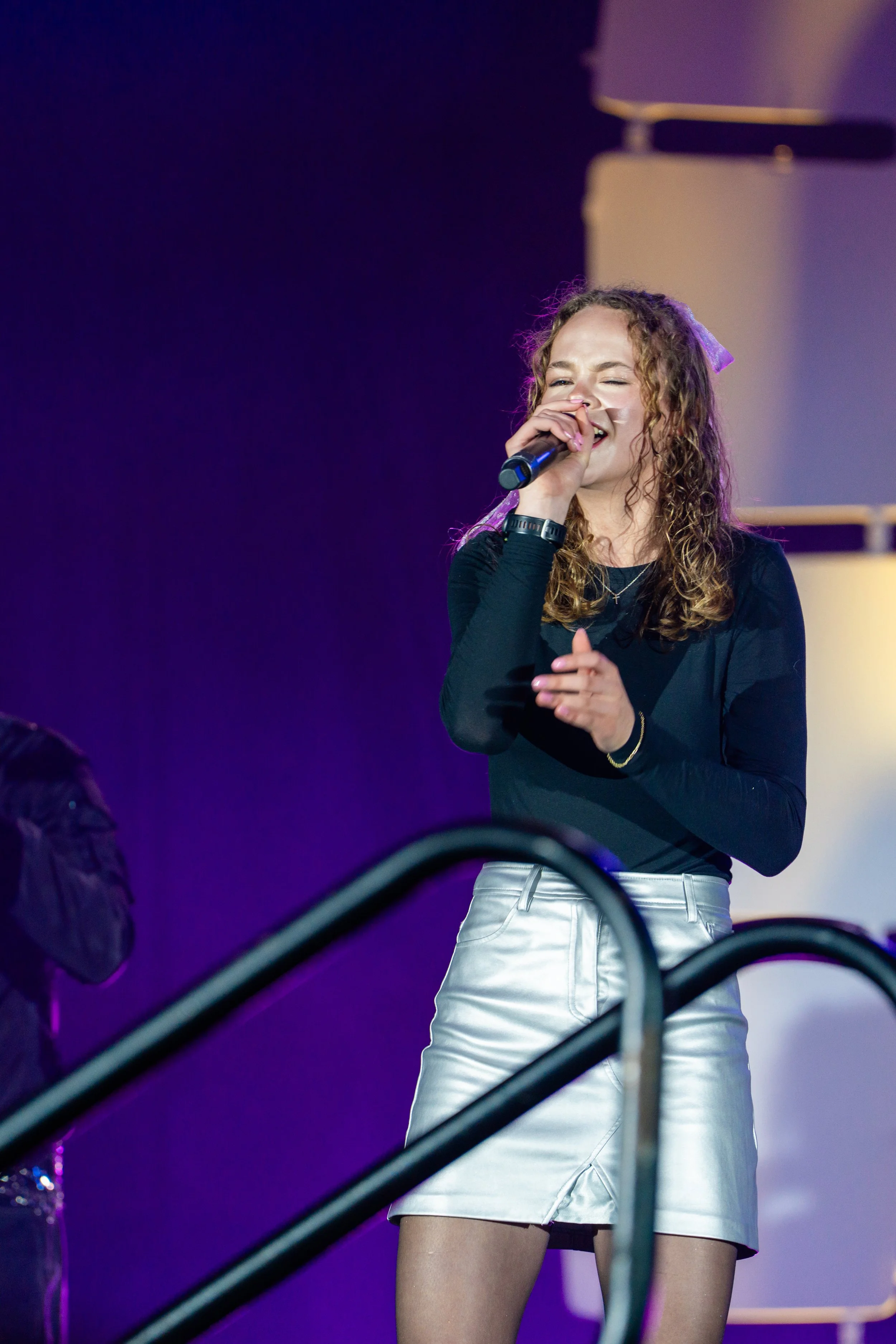 A young woman with curly hair singing into a microphone on stage, wearing a black top and white shiny shorts, with purple and yellow stage lighting.