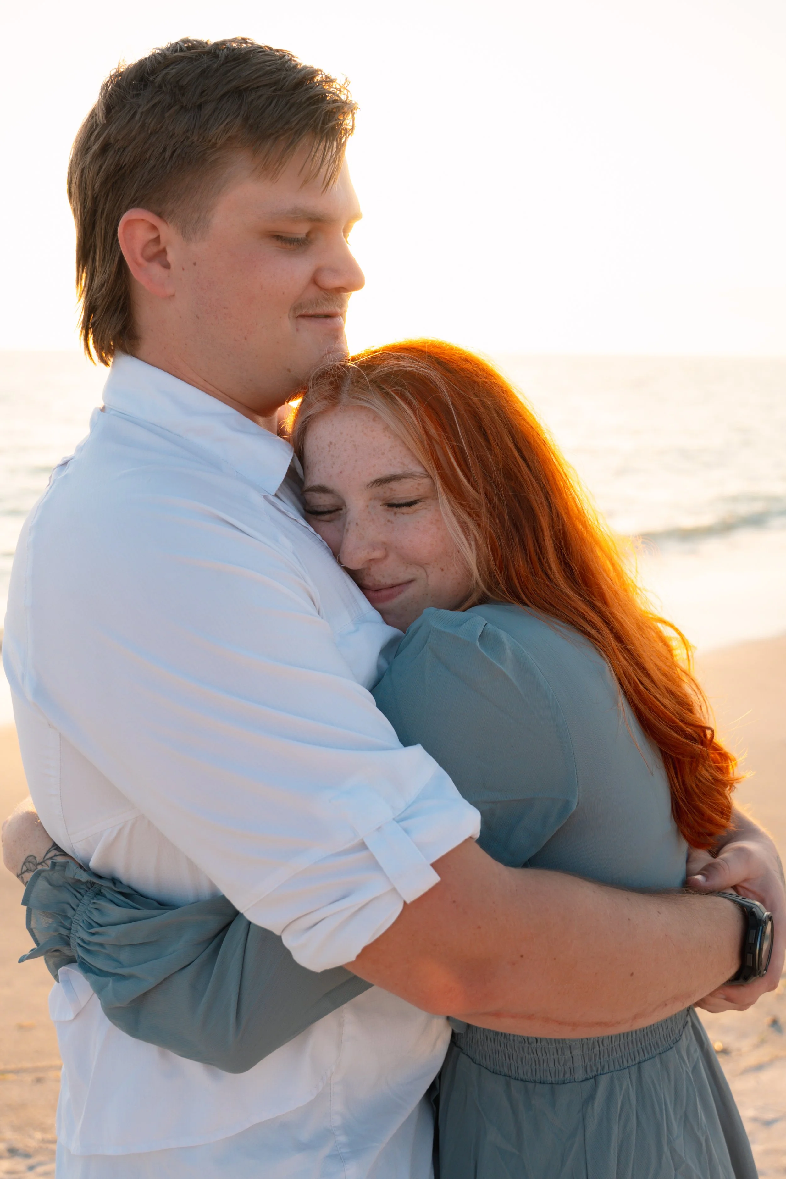 A couple hugging on a beach at sunset, with the woman smiling and the man gently holding her.