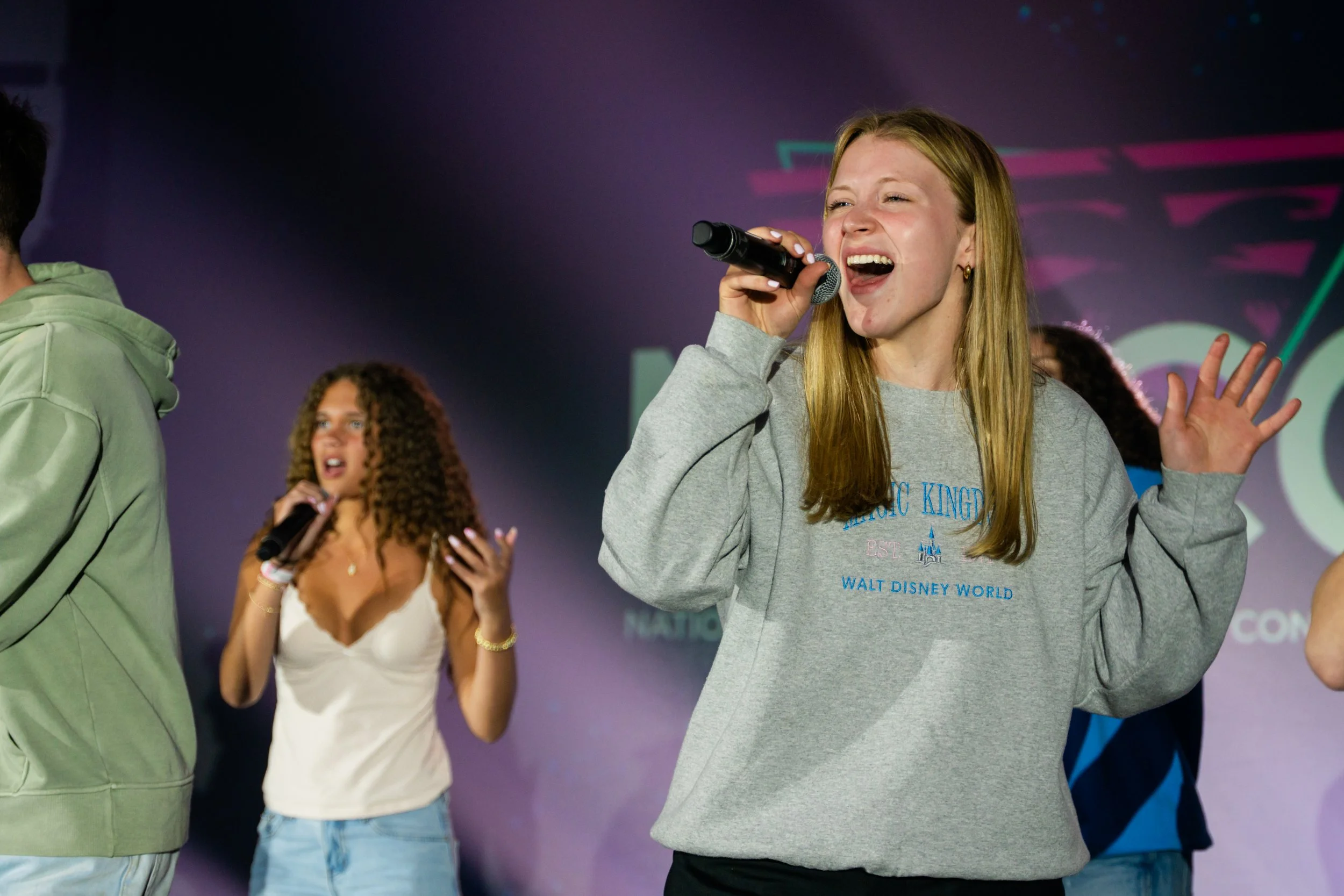 Group of young people singing and dancing on stage at Disney event, with girl in focus holding microphone and smiling.
