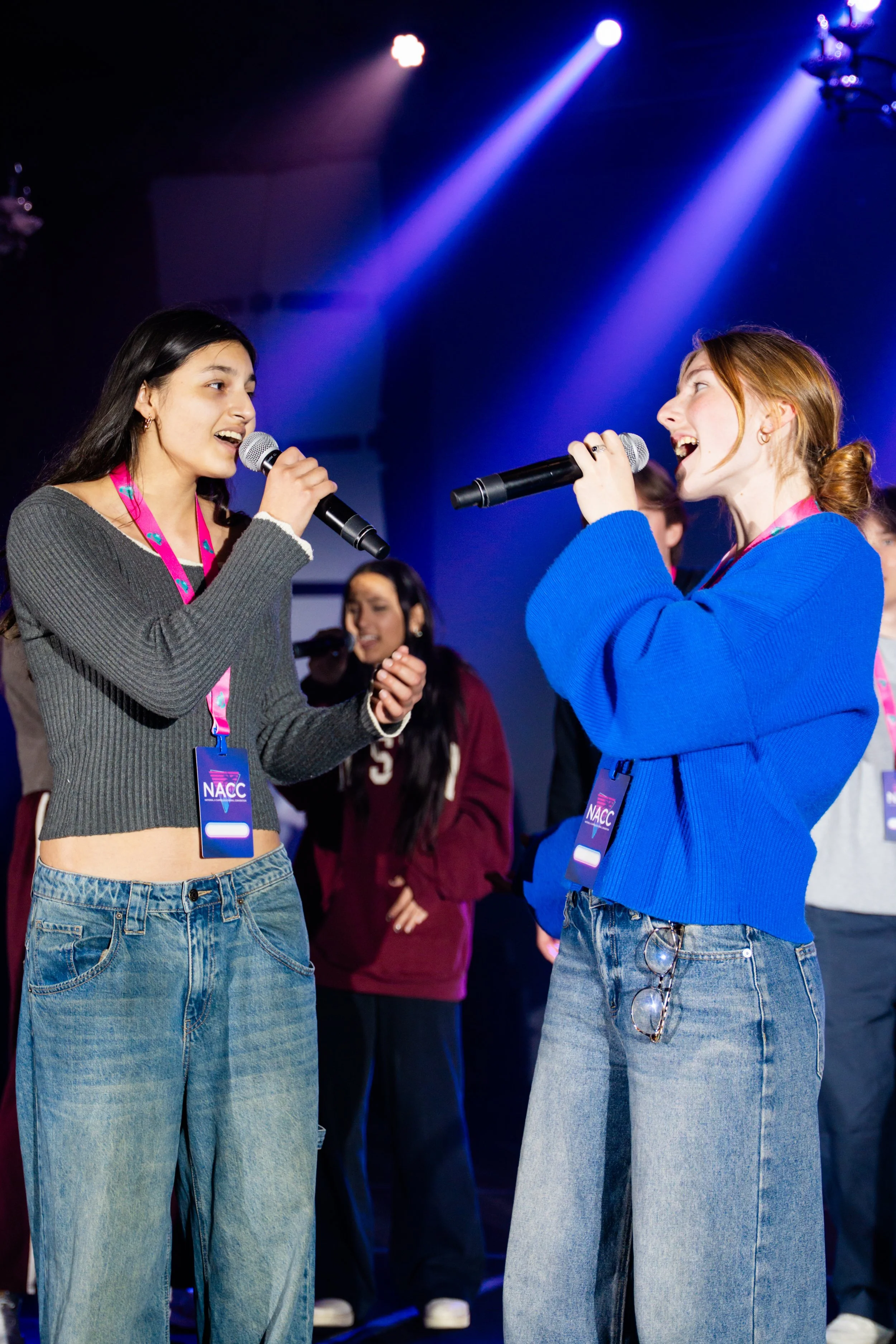 Two young women holding microphones and singing on stage with blue spotlights, wearing casual jeans and sweatshirts, with event badges around their necks.