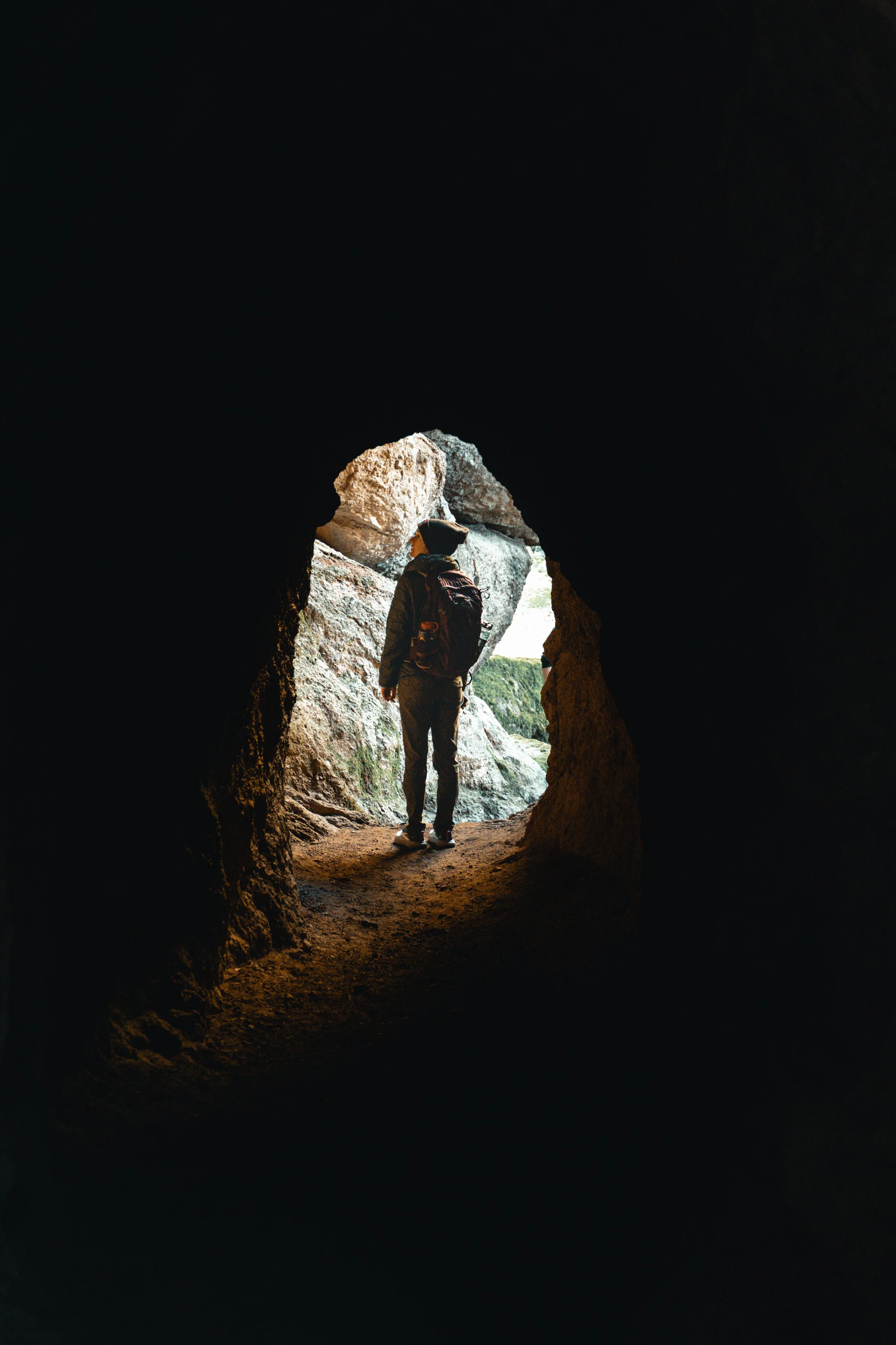 Person with a backpack standing at the entrance of a cave, looking outside at a rocky landscape.