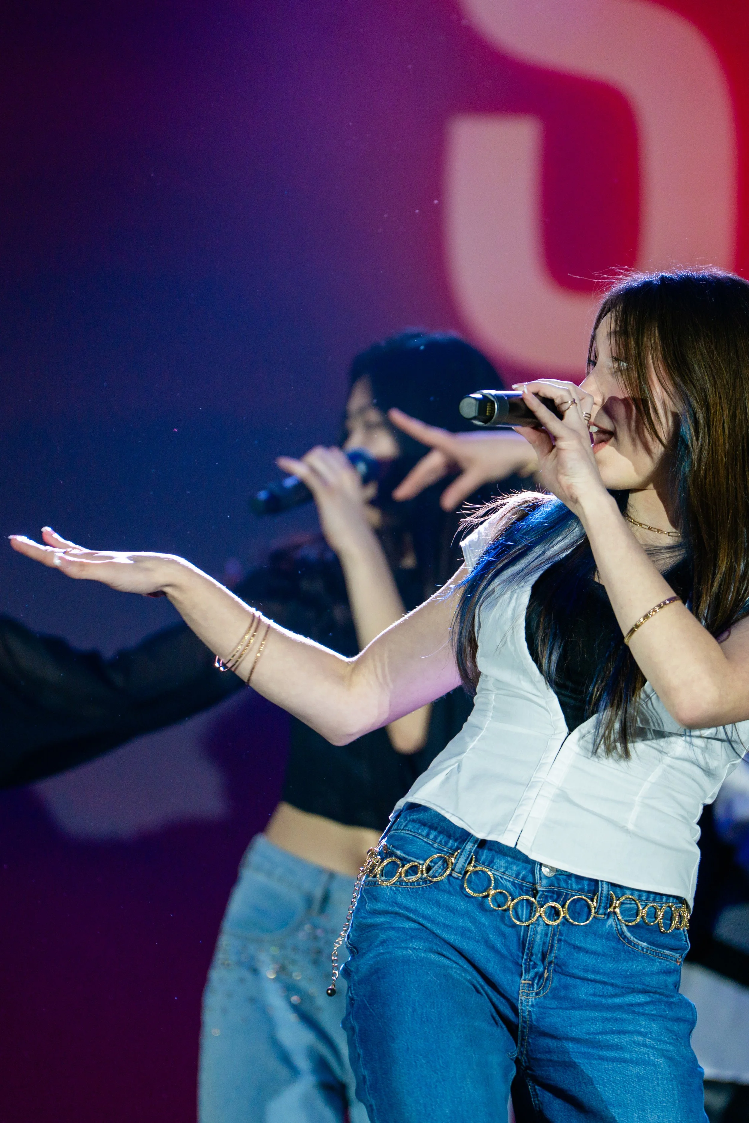 Female performer singing into a microphone on stage, wearing a white top and blue jeans with a decorative gold chain belt, with a blurred background featuring red, pink, and purple lighting.