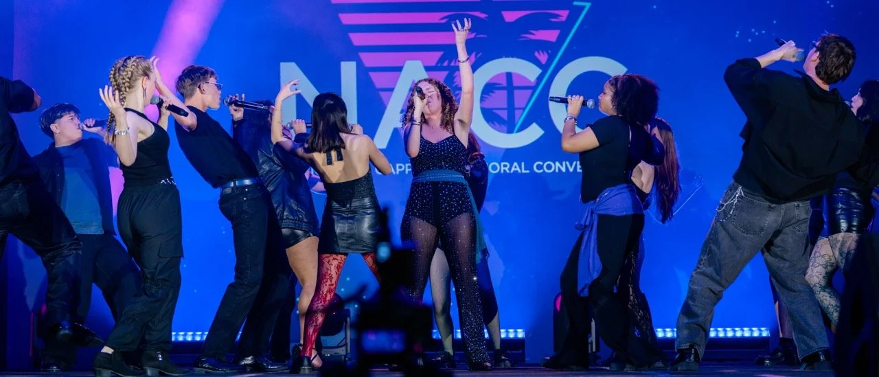Group of performers on stage holding microphones, singing, and dancing at the NACC conference, with a blue and purple background.