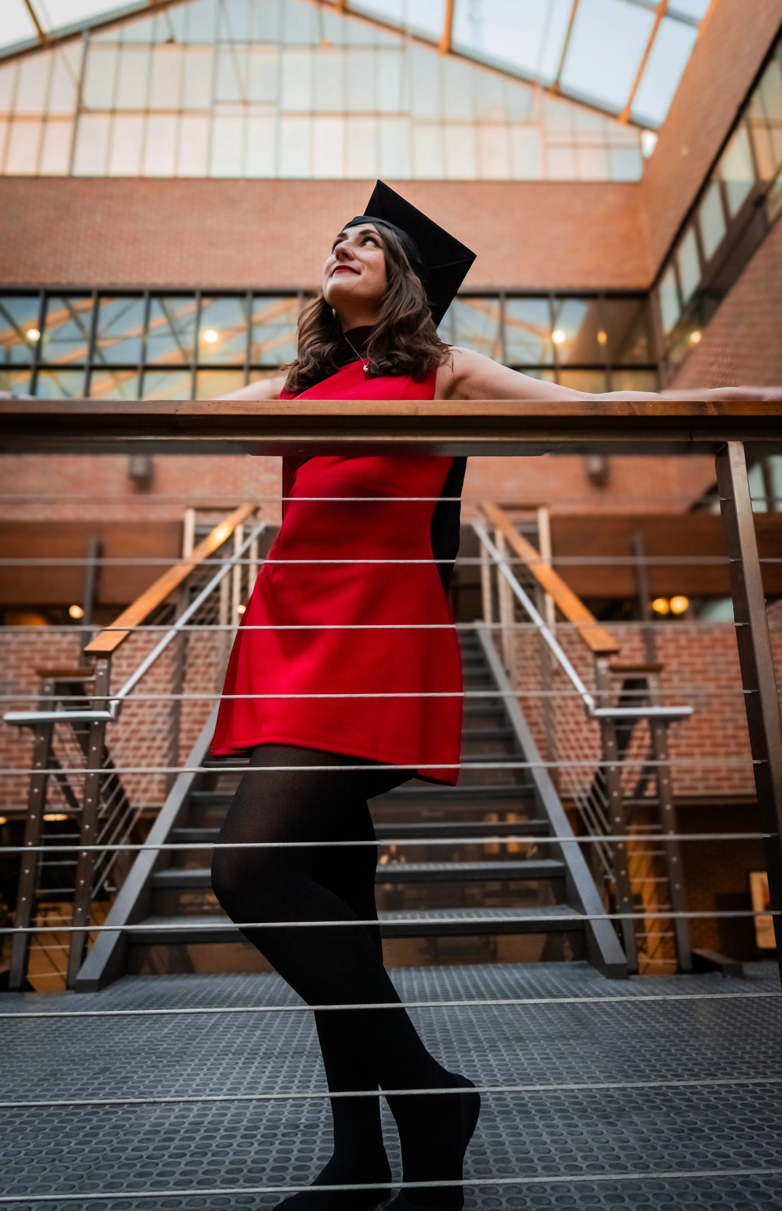 A young woman in a red dress and black tights, wearing a graduation cap, stands on a metal staircase inside a building with brick walls and glass windows, celebrating her graduation.