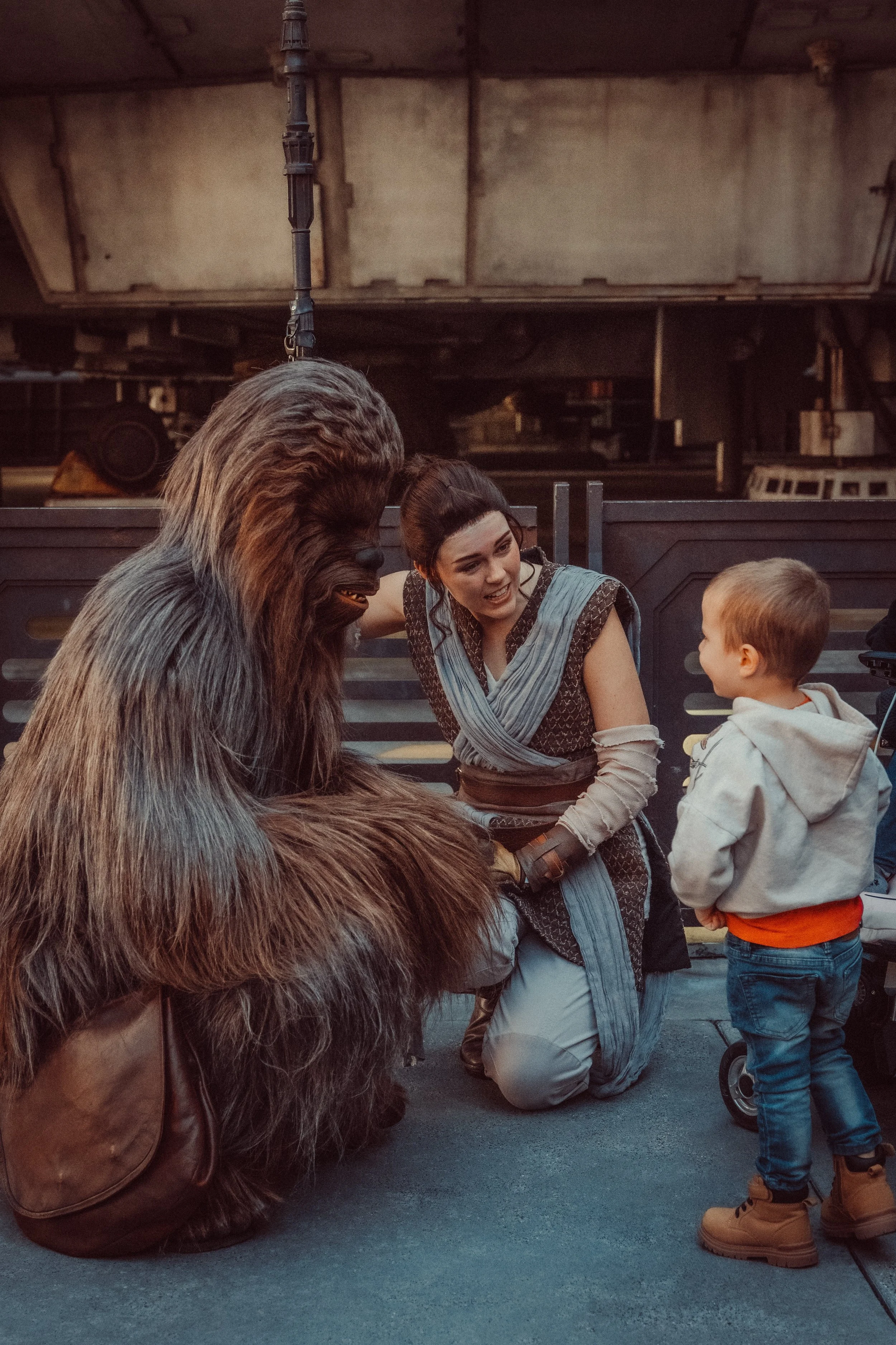 Person dressed as Chewbacca from Star Wars kneeling on the ground, having a conversation with a young boy and a woman in a costume resembling Princess Leia, in an industrial or outdoor setting.