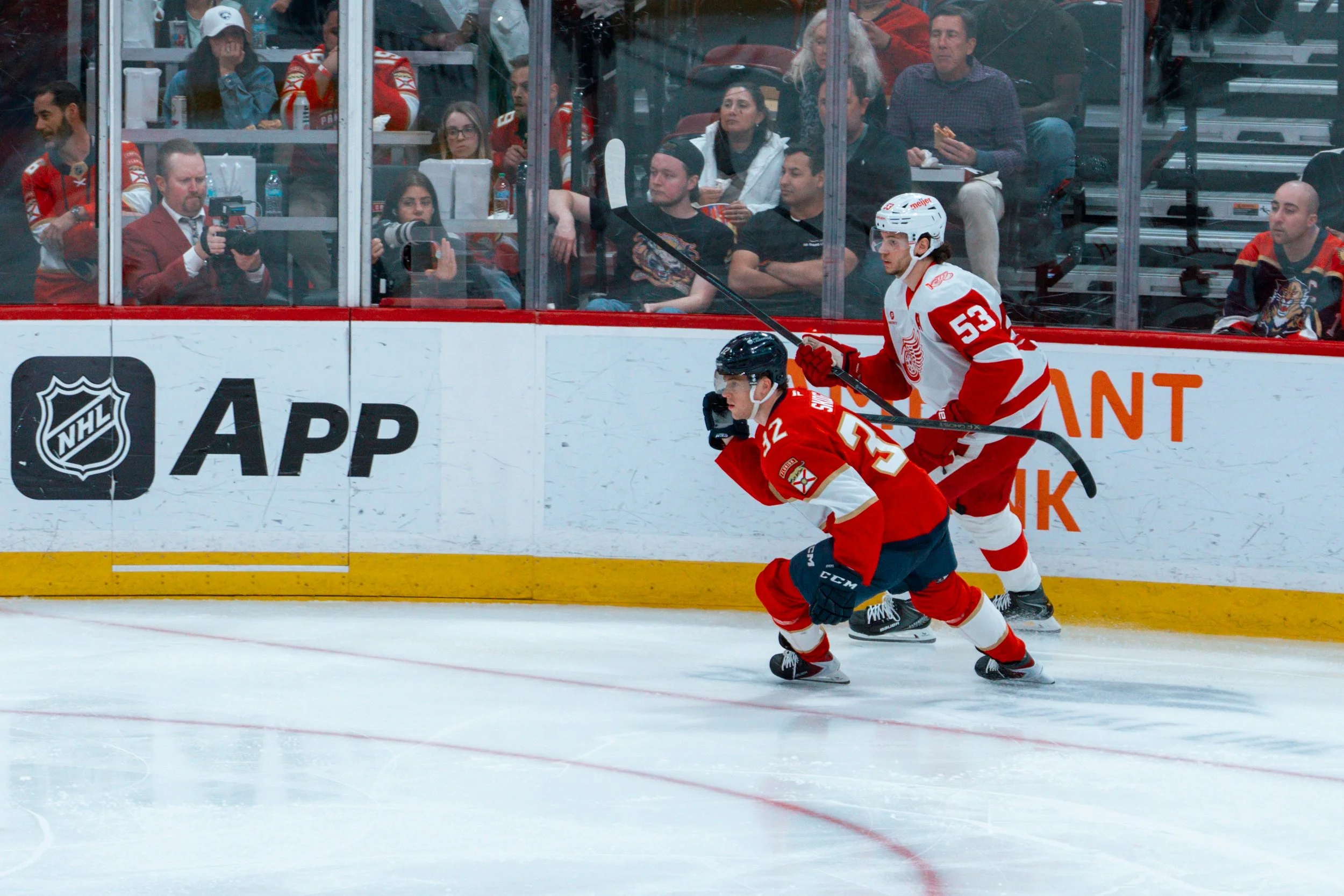 Hockey players in red and white jerseys skating on the ice rink during a game, with spectators watching from behind the glass.