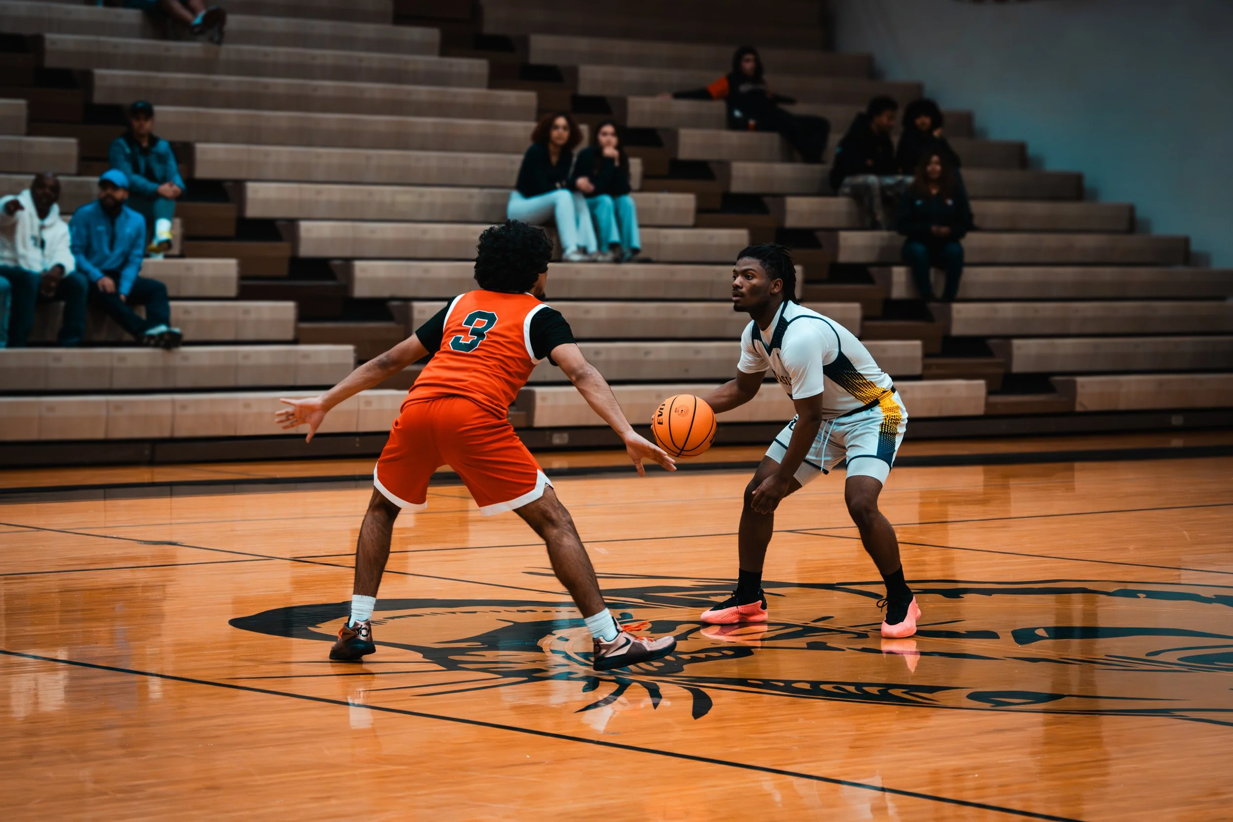 Two young men playing basketball on an indoor court, one in an orange jersey with number 3, defending, and the other in a white jersey with black, yellow, and blue accents, dribbling the ball. Spectators sit on wooden bleachers in the background.