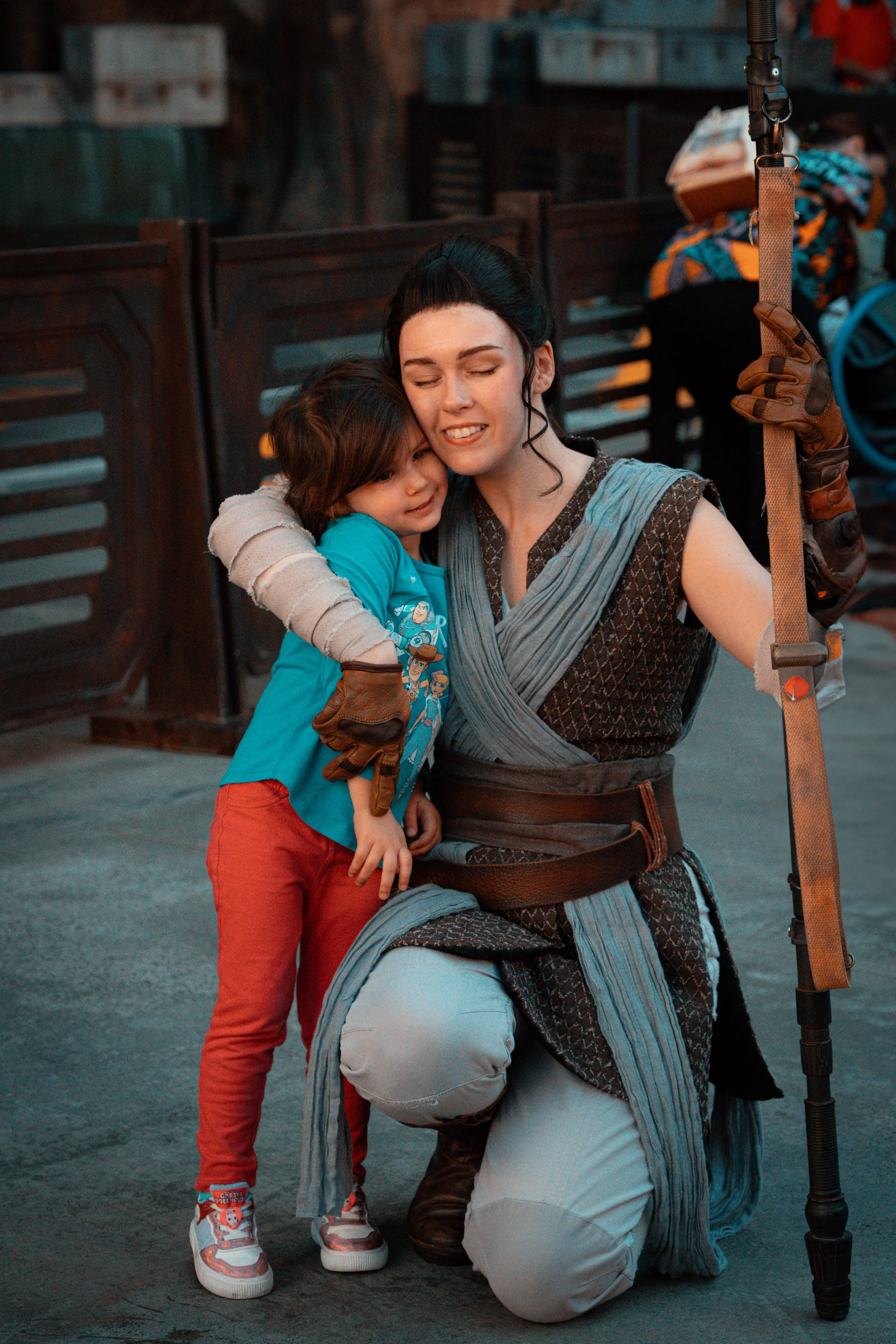 A woman dressed as a Jedi kneels on one knee, hugging a young boy dressed as a Jedi. They are indoors, with a rustic background, and appear to be sharing an emotional moment.