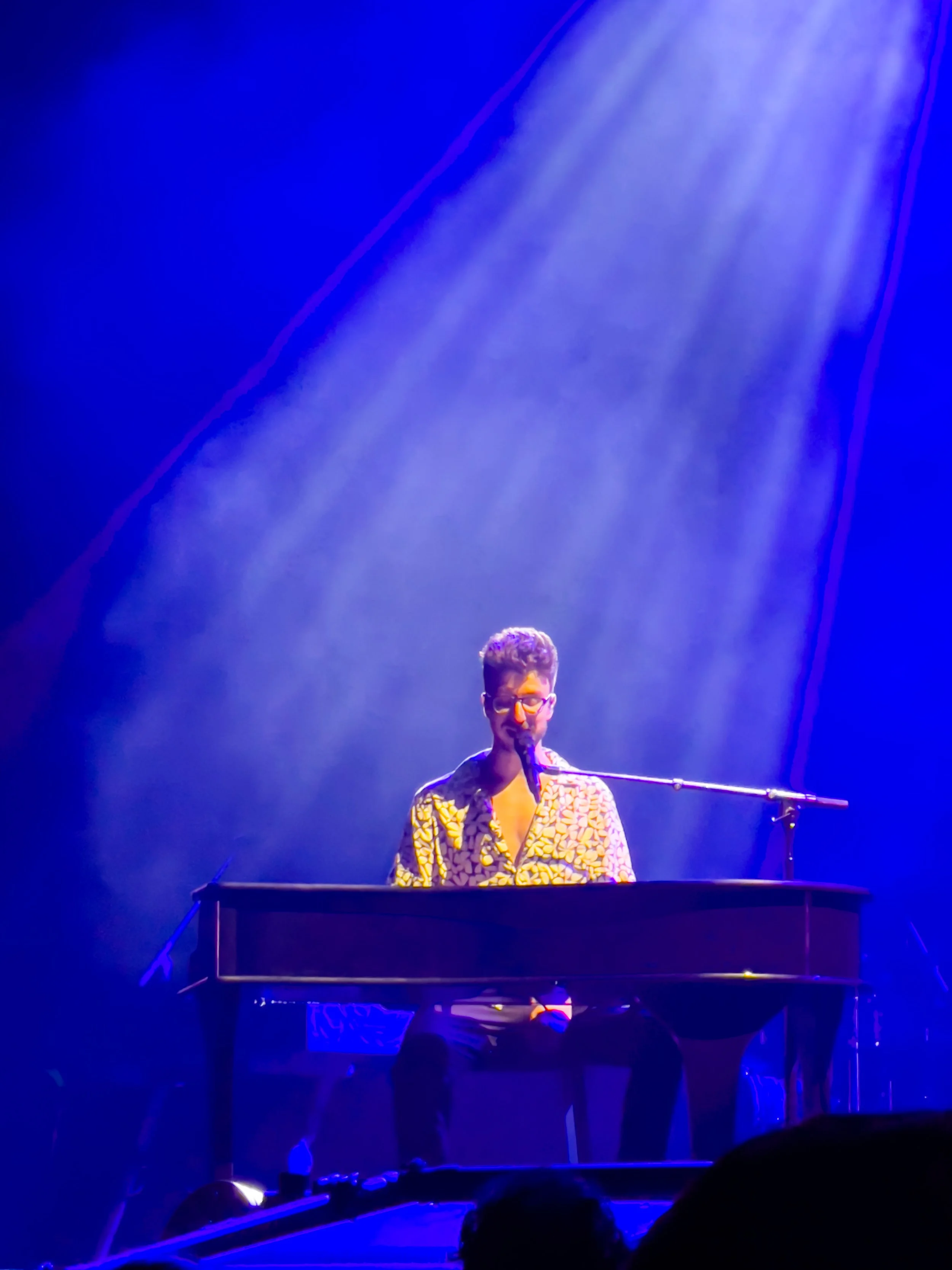 A man wearing glasses and a patterned shirt is sitting at a piano on a stage, singing into a microphone, with blue stage lighting and a beam of light shining down on him.
