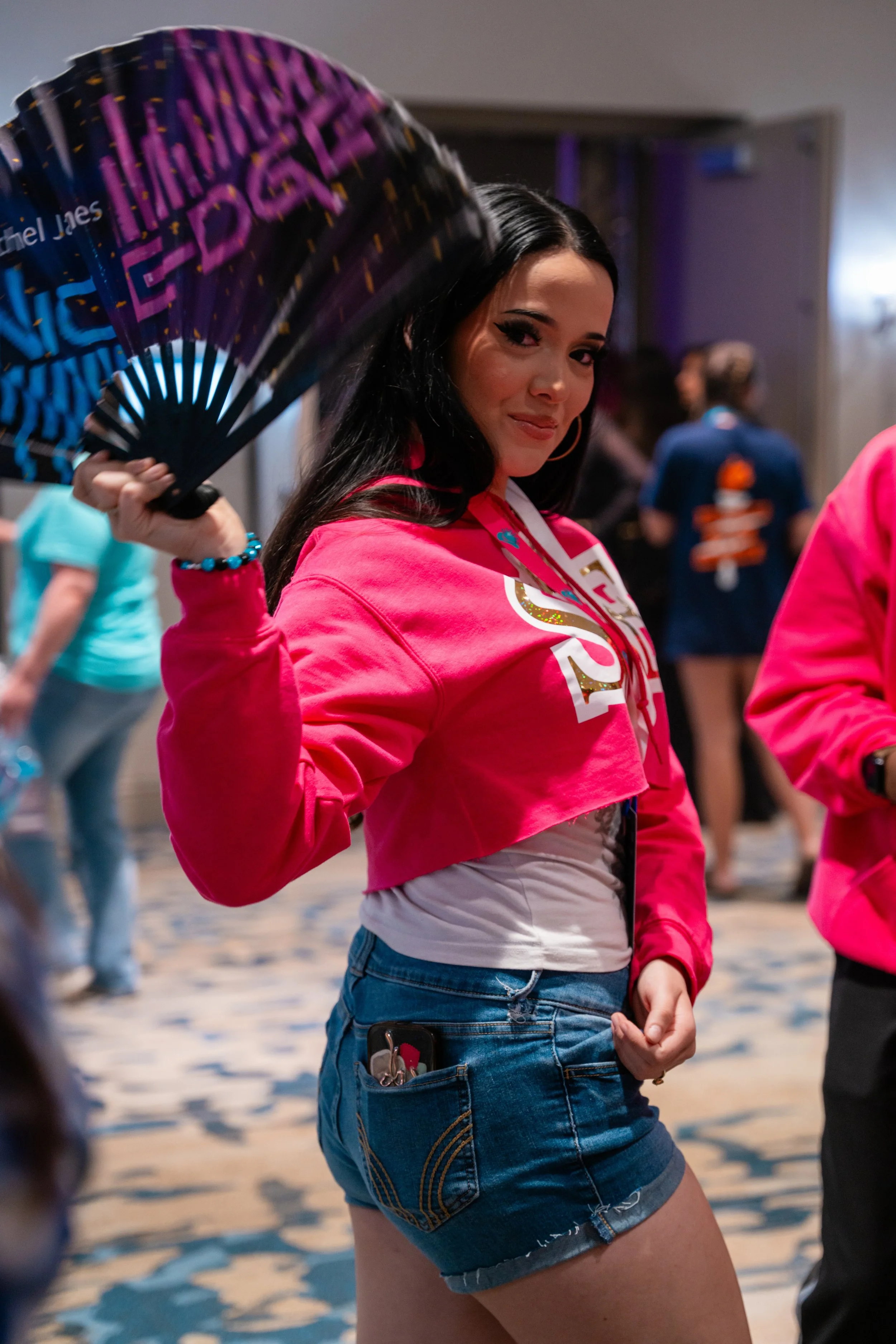 A young woman with long black hair, wearing a pink cropped hoodie and denim shorts, holding a black and purple fan with Gold accents, standing in a crowded indoor space.