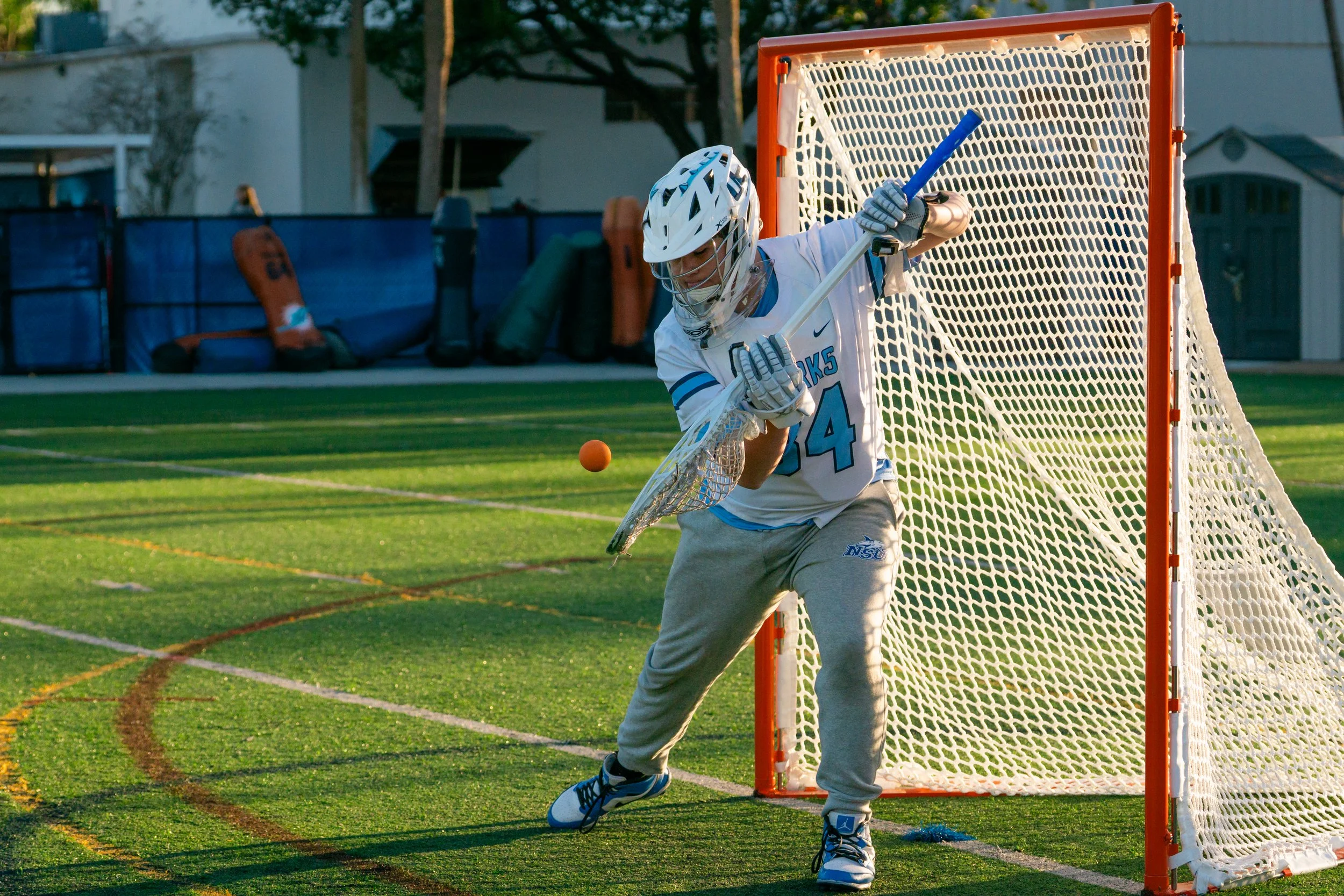 A lacrosse goalie in gear, including a helmet and gloves, attempting to block a shot near the goal during a game outdoors on a grassy field.