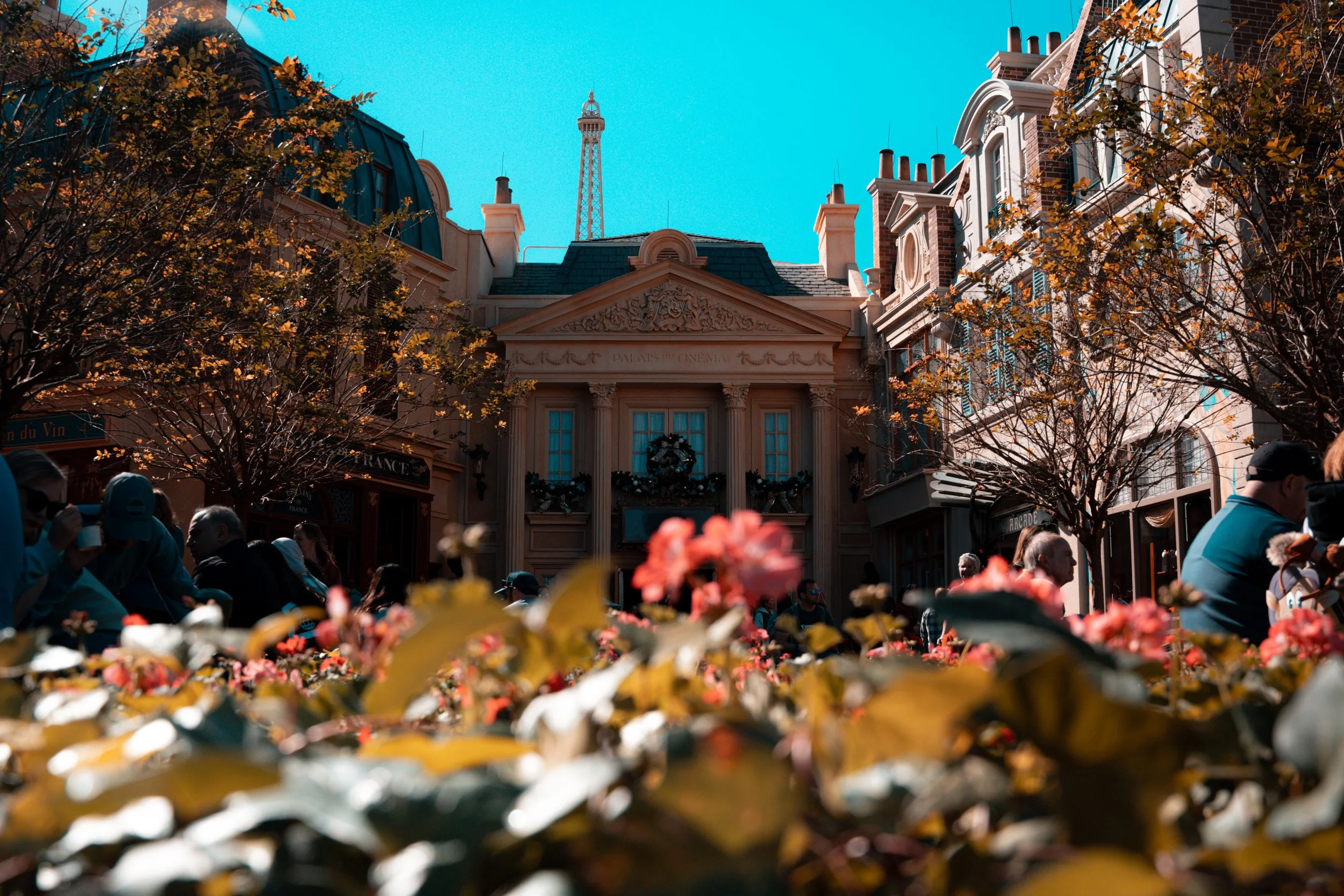Crowd of people in a shopping area with European-style architecture, including a building with columns, wreaths, and a decorative facade, with a tall tower in the background and trees with autumn leaves.