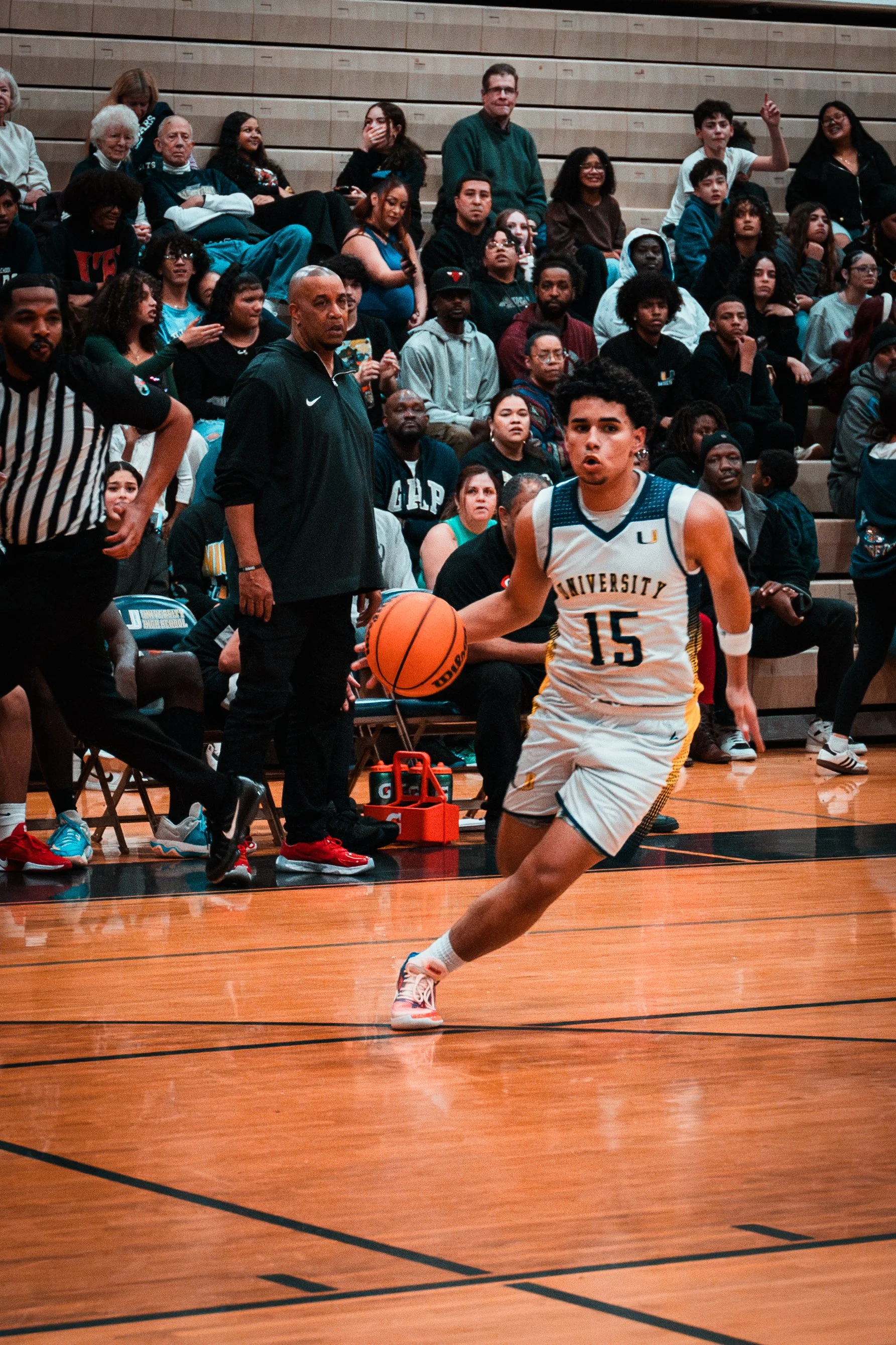 A basketball player wearing jersey number 15 for University is running on the court during a game, with an audience watching from the bleachers in the background.