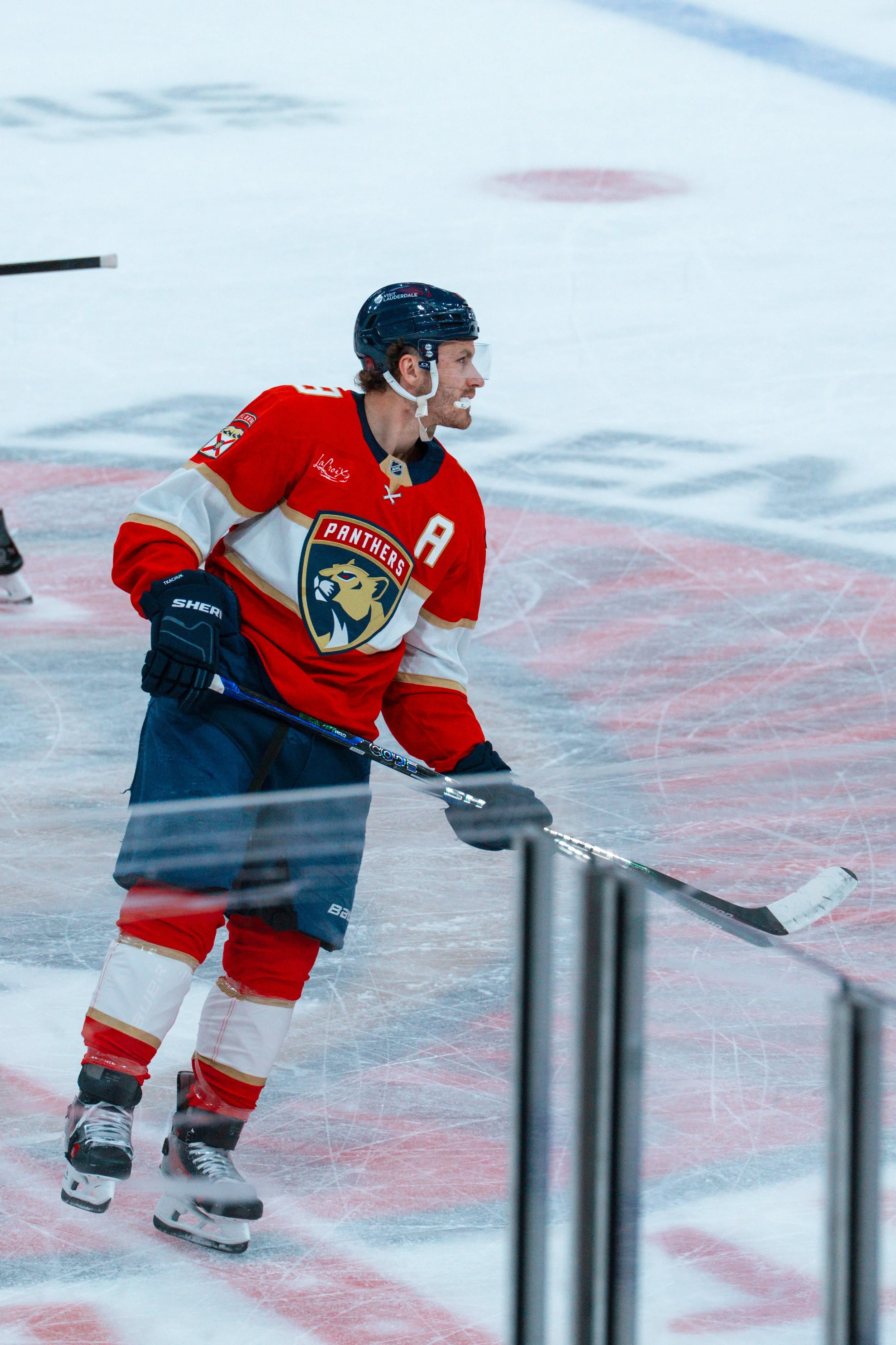 An ice hockey player in a red and white uniform with a black helmet, holding a hockey stick on the ice rink.