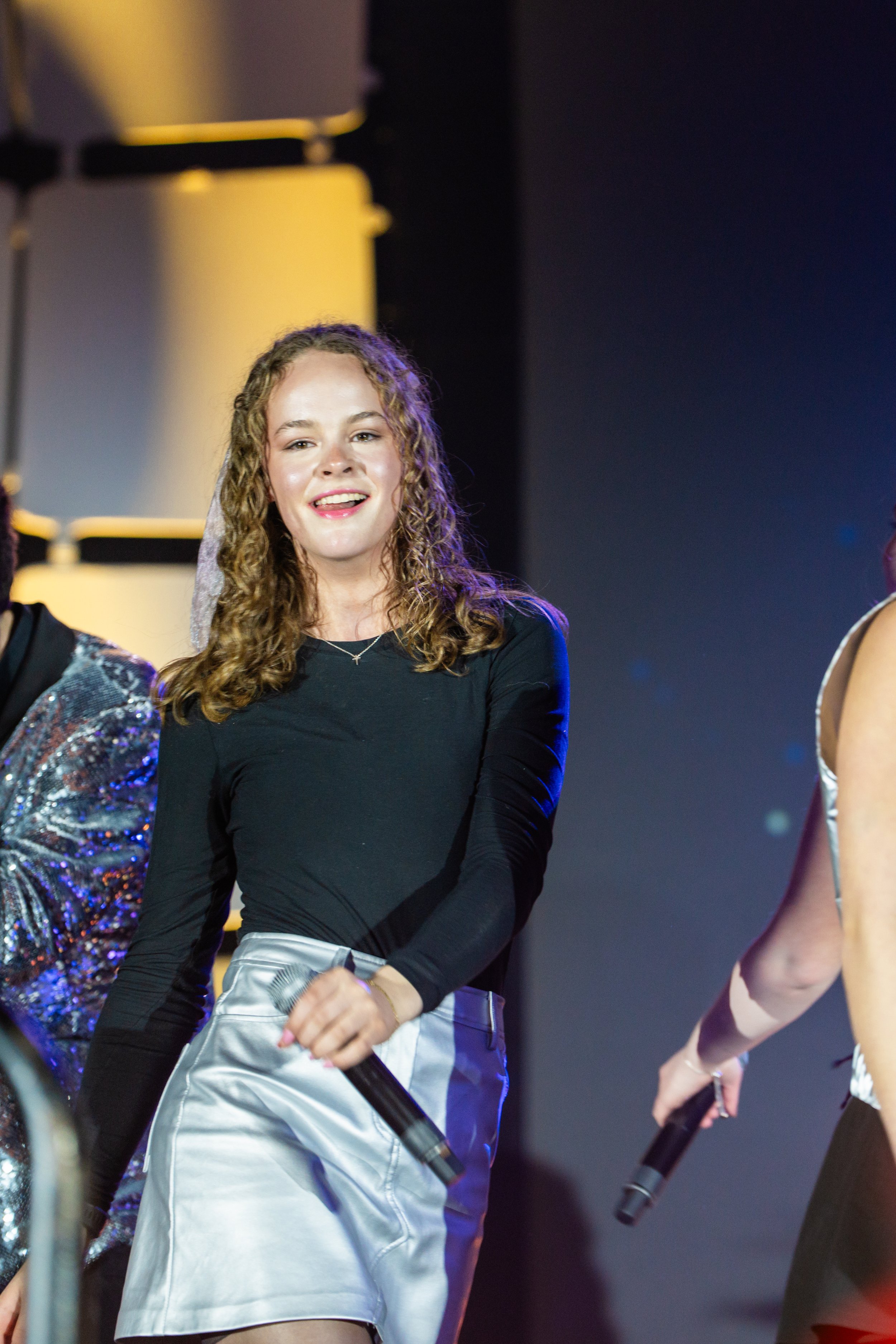 A young woman with curly blonde hair smiling and holding a microphone on stage during an event, wearing a black top and shiny silver skirt.