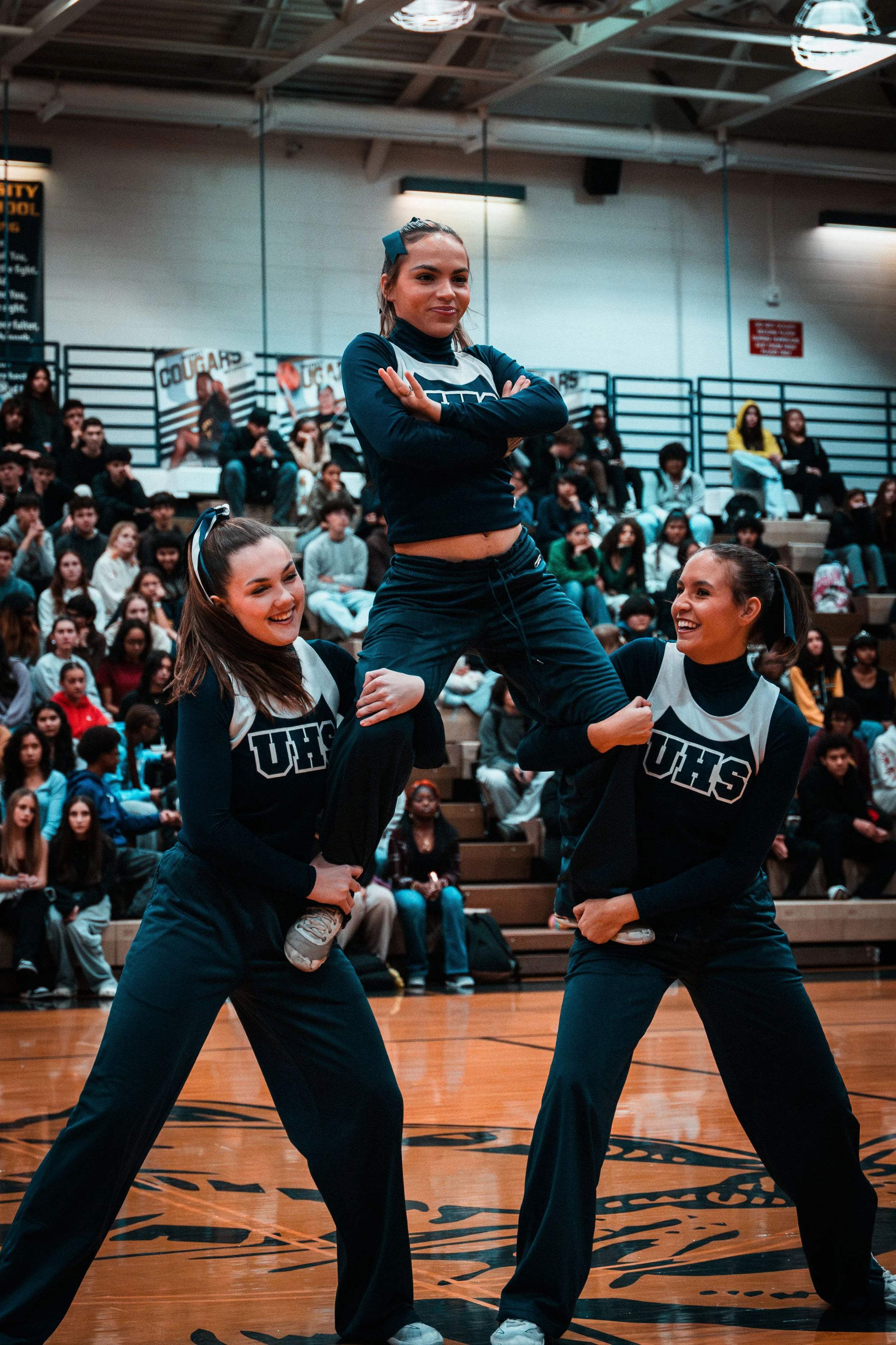 Three cheerleaders perform a stunt on a gymnasium court in front of a crowd of students, with two cheerleaders holding up a third in the middle.
