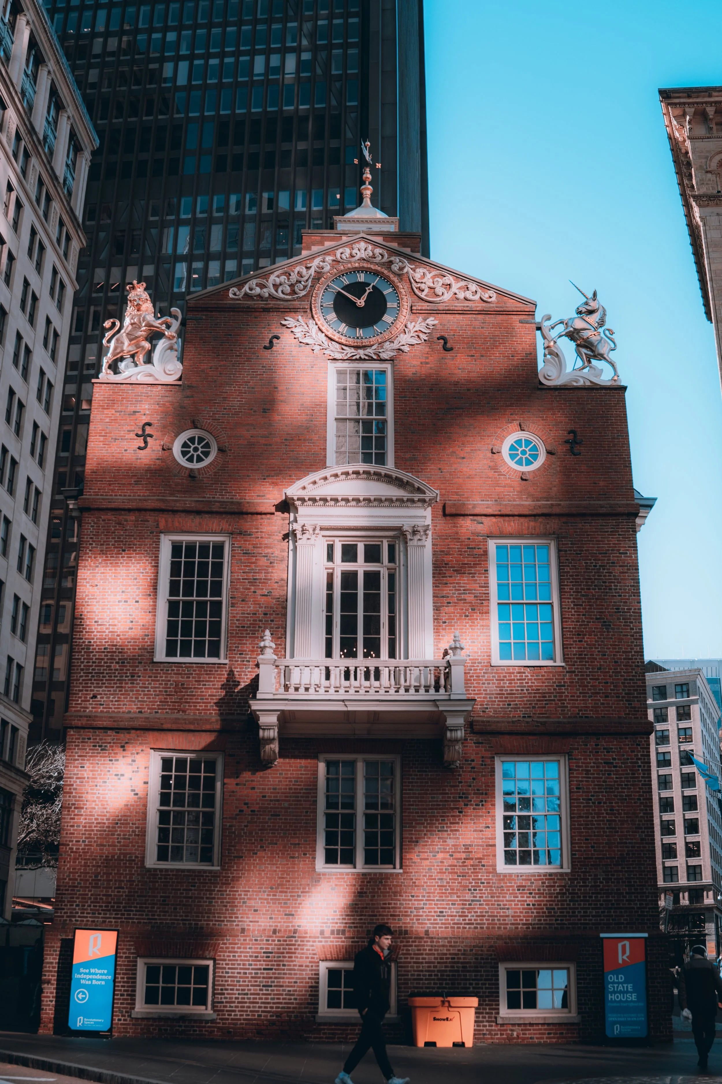 A historic red brick building with a clock and decorative statues on the roof, located in an urban area with modern skyscrapers. The building has several windows, a small balcony, and shadows cast across its facade.